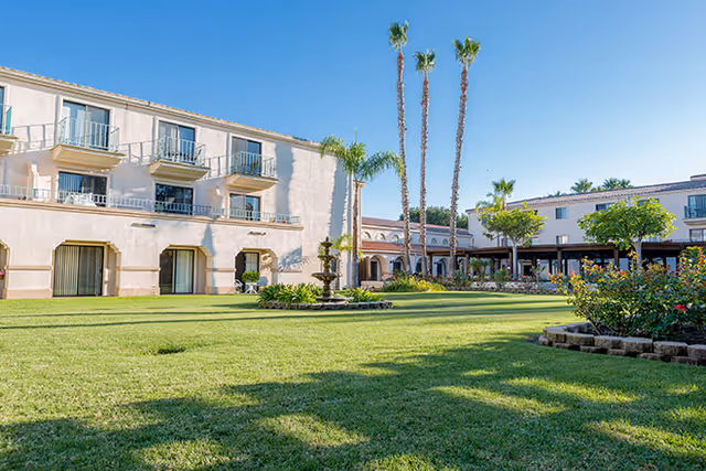 Two-story tan senior living building with balconies, palm trees, and a grassy courtyard.