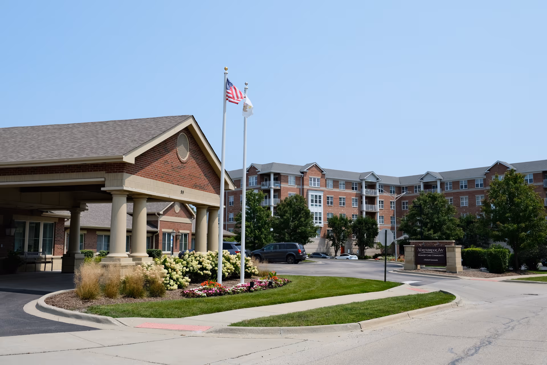 Front entrance of Northbrook Inn Memory Care Community showing a covered porte-cochere, flagpoles, landscaped flowerbeds, and the multi-story brick building behind.