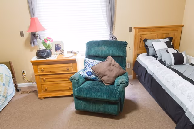 A cozy senior living bedroom featuring a wooden bed with a black, white, and gray patterned comforter and multiple pillows. Next to the bed is a wooden nightstand with a pink lamp, a framed photo, a small plant, and a radio. In front of the nightstand is a green upholstered armchair with two decorative pillows. The room has beige walls and carpeted flooring, with a window covered by white blinds and gray curtains.
