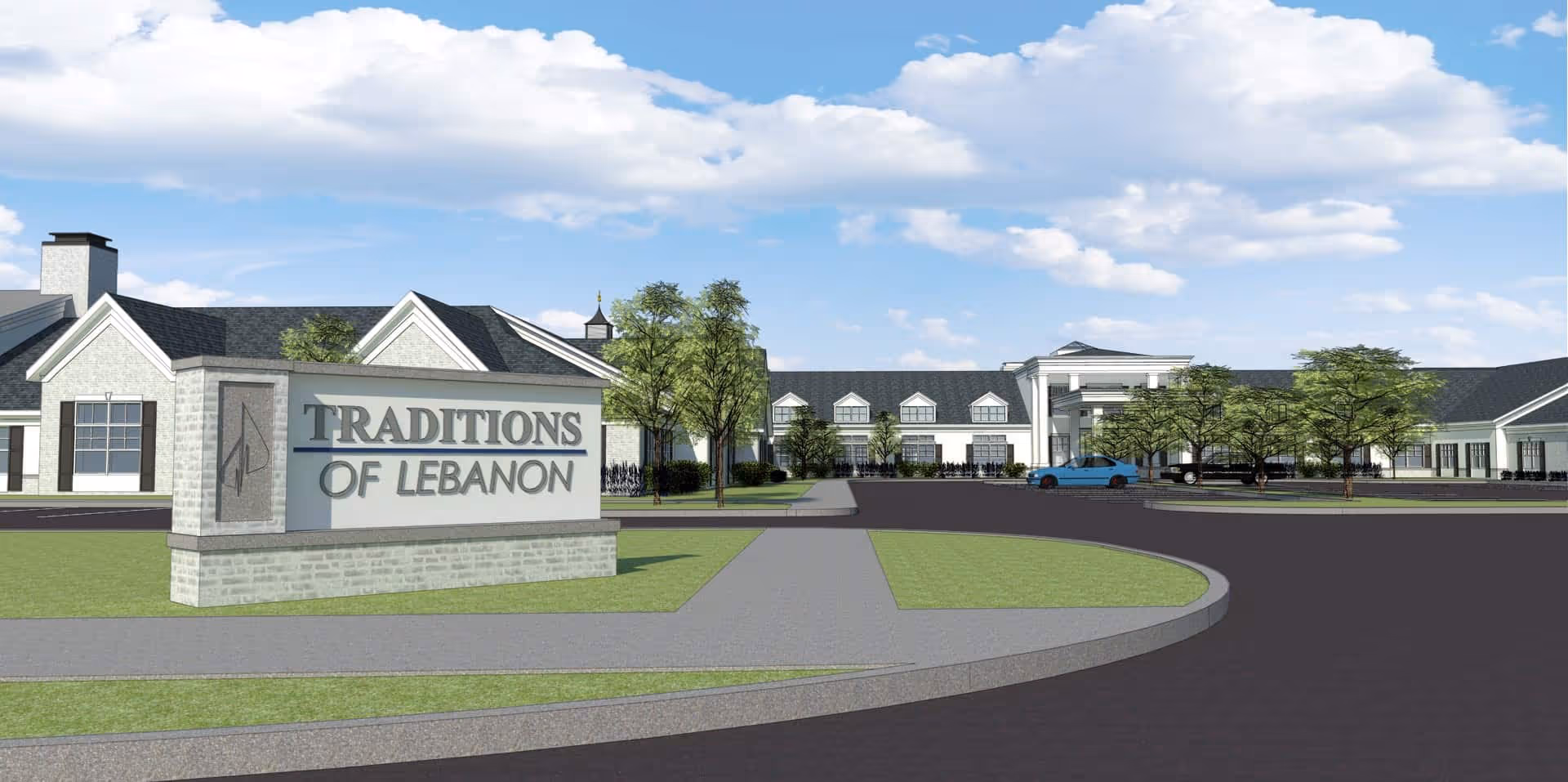 Exterior view of the Traditions of Lebanon senior living facility with a large sign in the foreground displaying the facility's name. The building has a traditional architectural style with white walls, dark roofing, and multiple windows. There are trees and a driveway leading to the entrance under a partly cloudy sky.