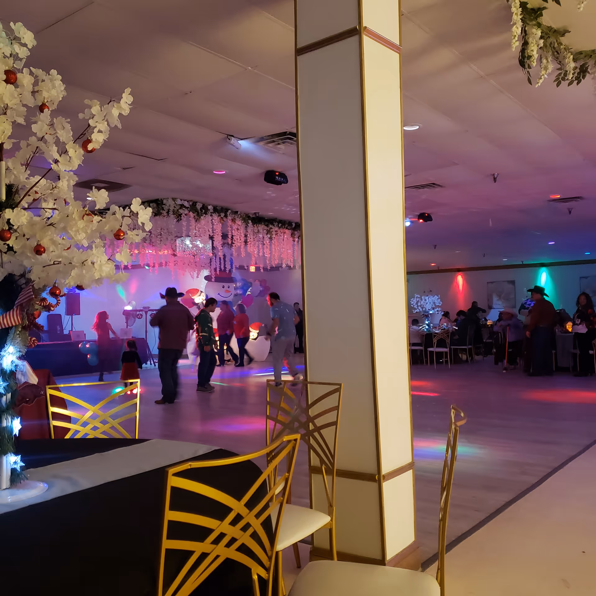 Indoor party hall with festive decor, a central pillar, people dancing, and round tables.