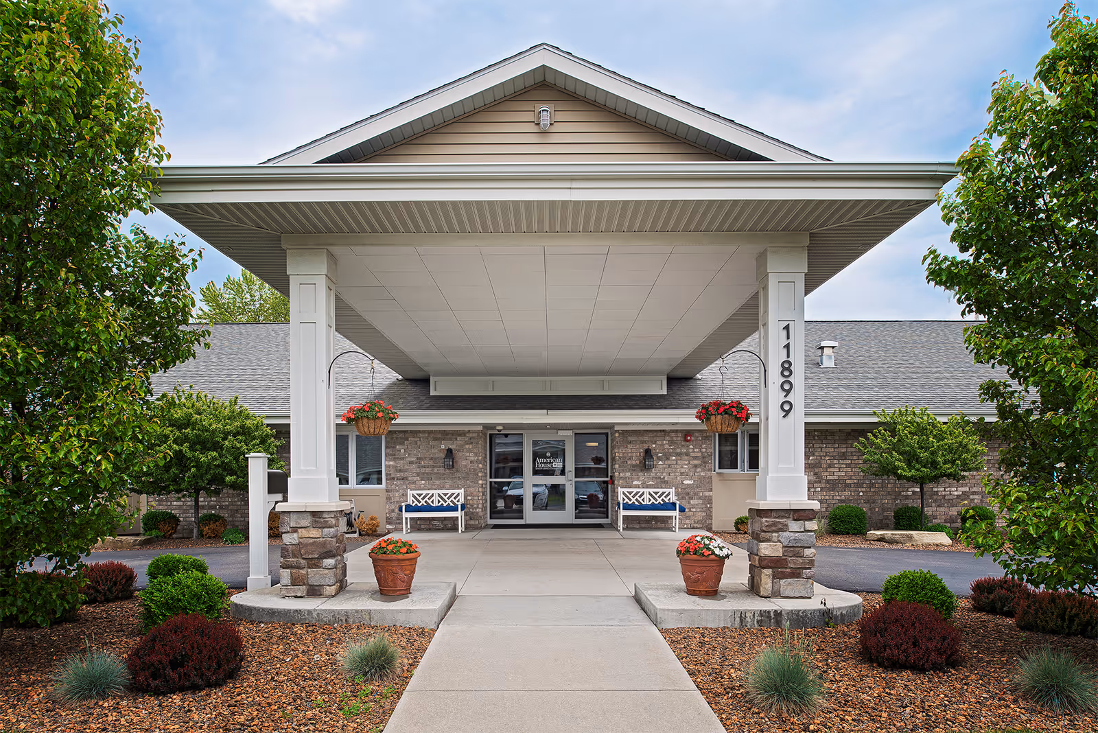 Covered entrance of the American House Holland facility with a porte-cochere, benches, potted plants, and the number 11899 on a column.