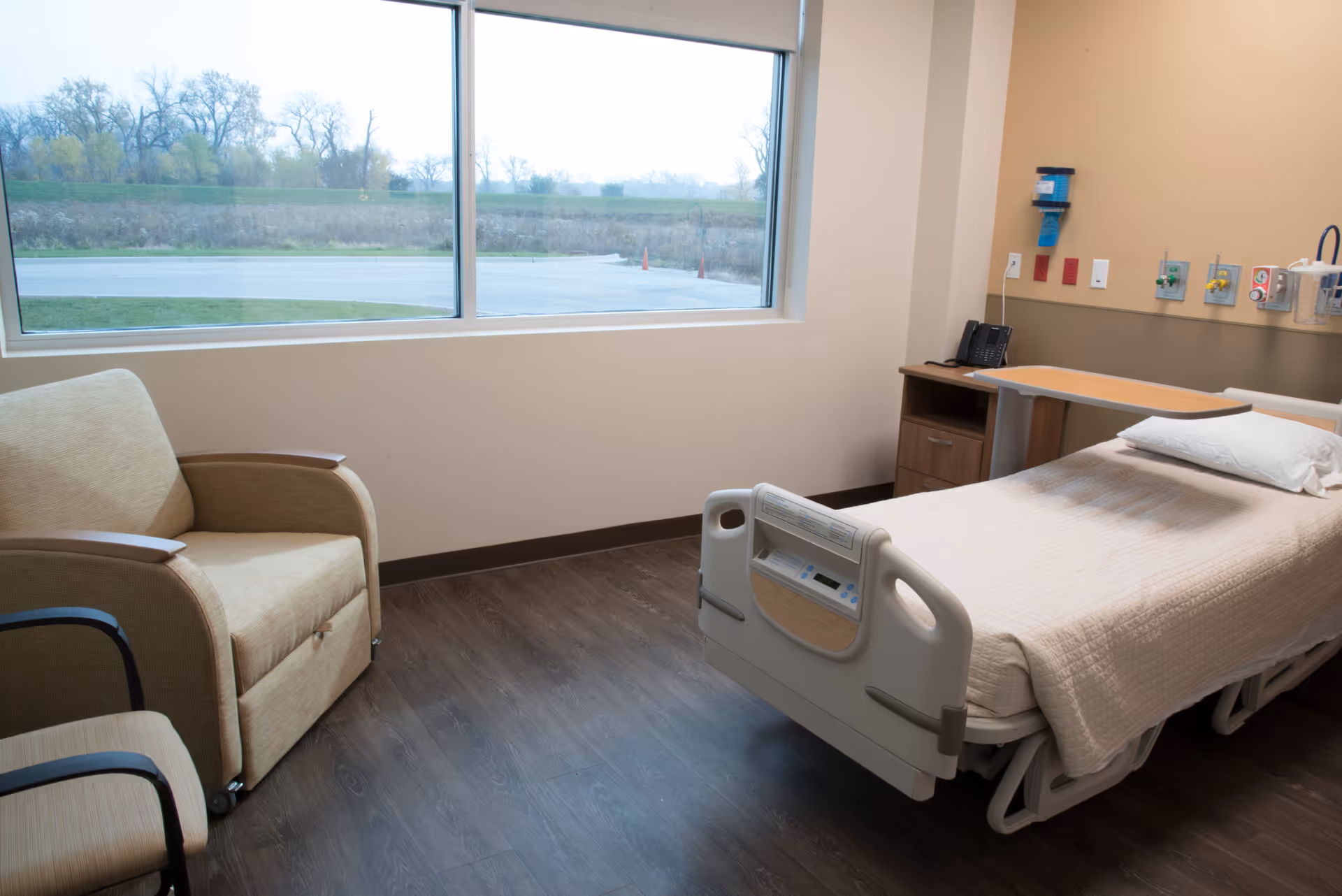 Patient room with hospital bed, chairs, and medical equipment by a large window.