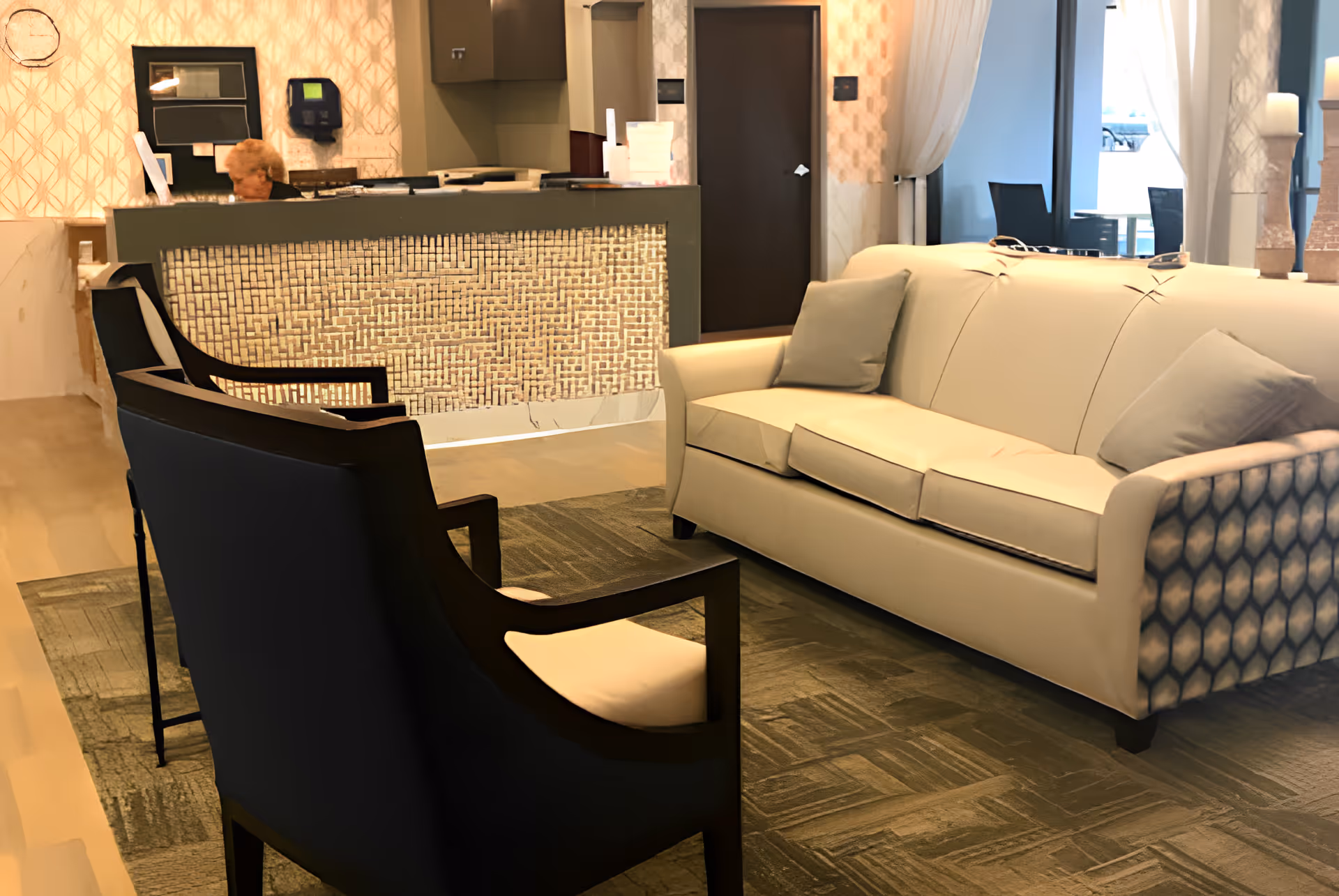 Reception/lobby area with a beige sofa, two dark armchairs, and a front desk with a person behind it.