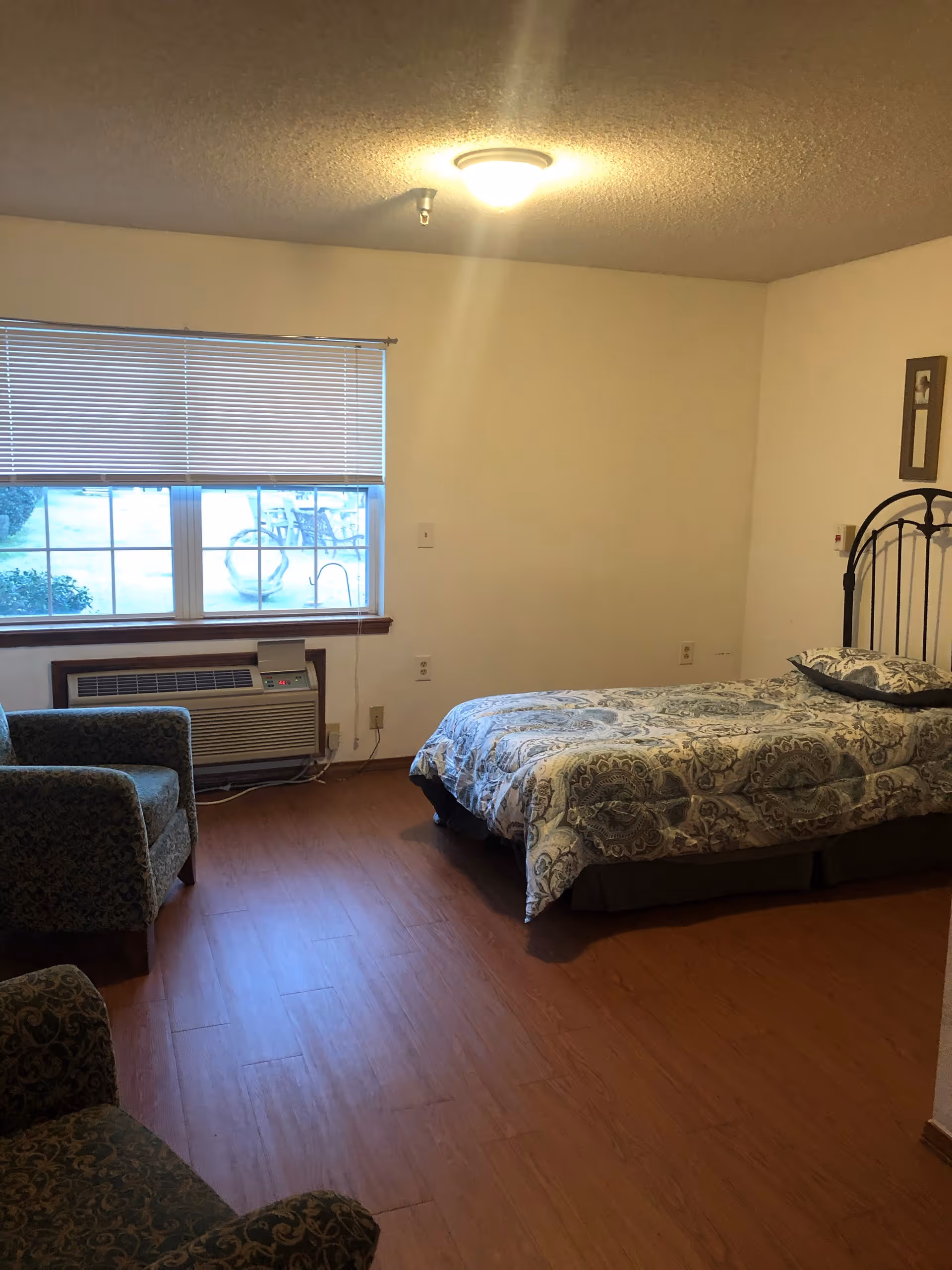 A simple bedroom with a single bed covered in a patterned comforter, two upholstered chairs with a floral design, a large window with closed blinds, and a wall-mounted air conditioning unit below the window. The room has wooden flooring and plain white walls with a ceiling light fixture.