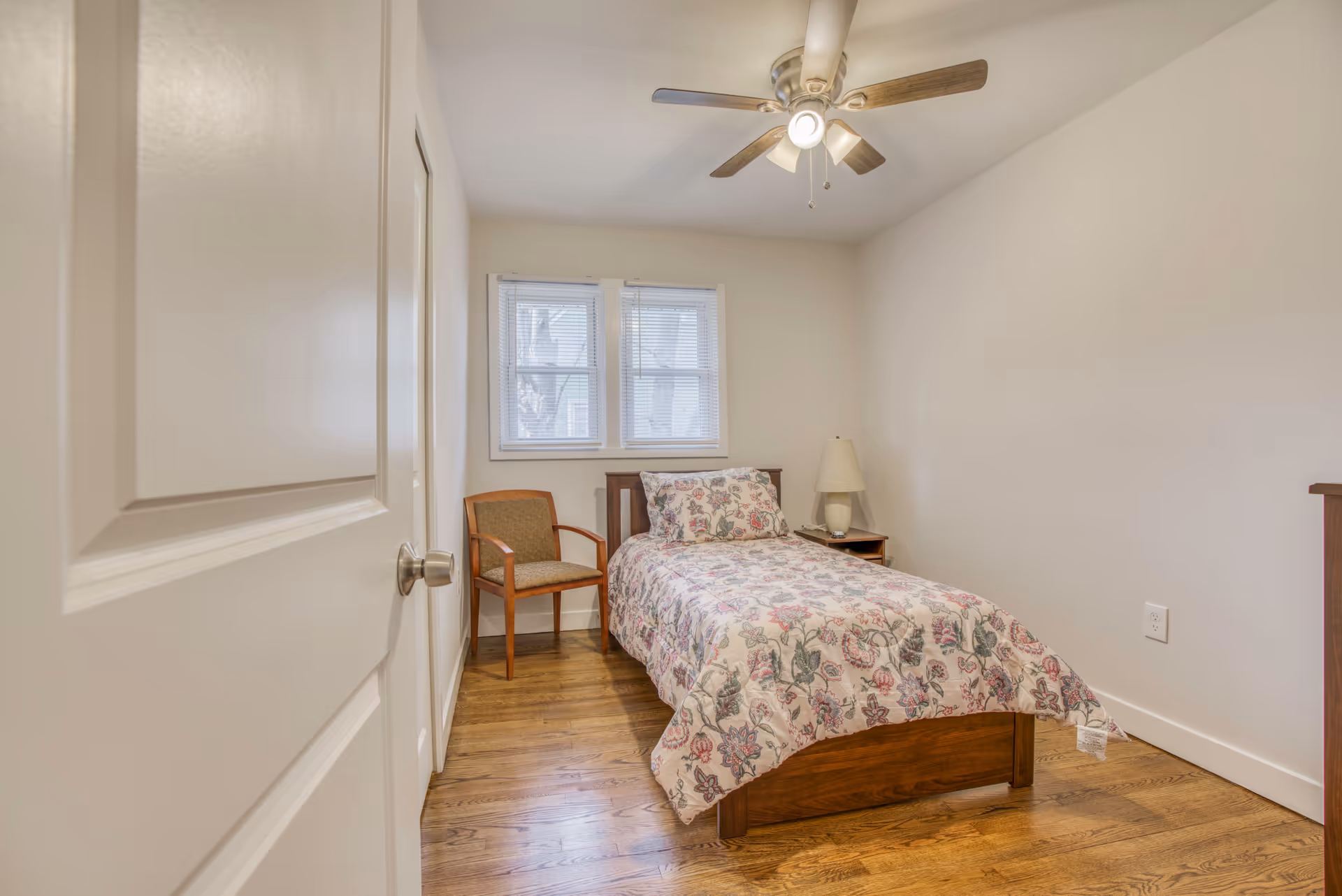 Small bedroom with single bed, floral bedspread, chair, nightstand, and ceiling fan.