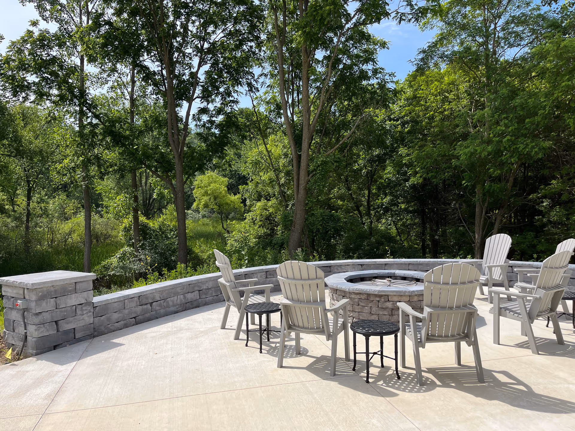 Outdoor patio area with a circular stone fire pit surrounded by several white Adirondack chairs and small black side tables. The patio is bordered by a low stone wall and is set against a backdrop of green trees and foliage under a clear blue sky.