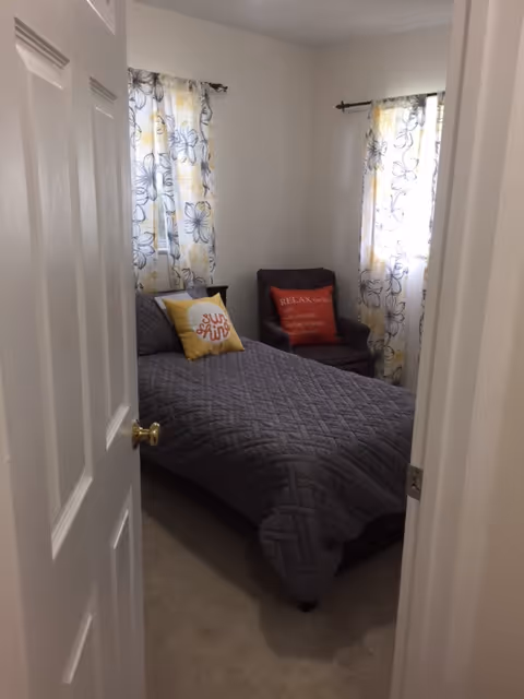 Small bedroom with a gray quilted bed, floral curtains, and a corner chair.