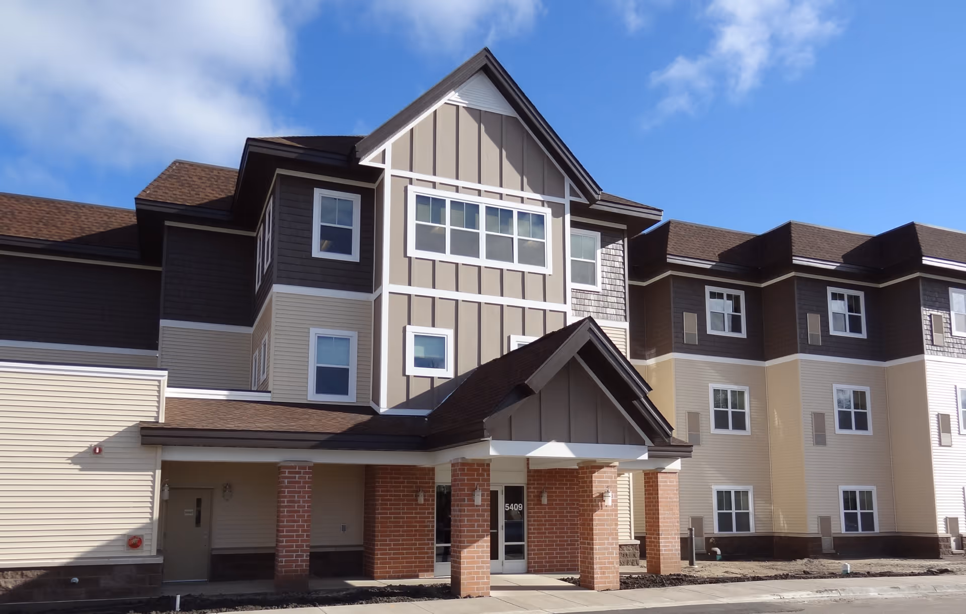 Exterior view of a multi-story senior living facility building with a covered entrance supported by brick columns under a blue sky with some clouds.