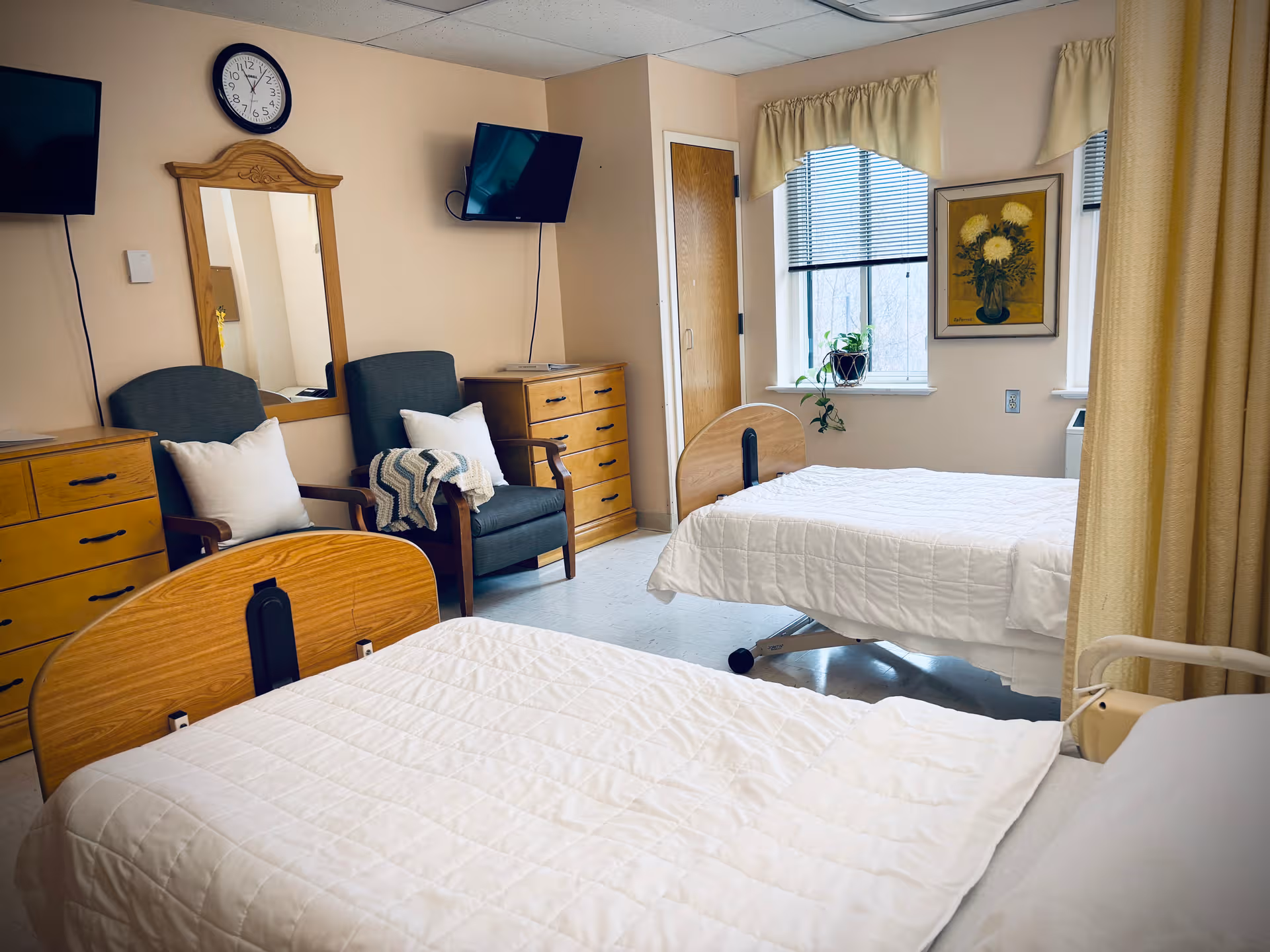 Two beds and seating in a shared senior living bedroom with dressers and a wall clock.