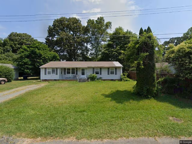 Single-story ranch-style house with a front porch set behind a large grassy yard and trees.