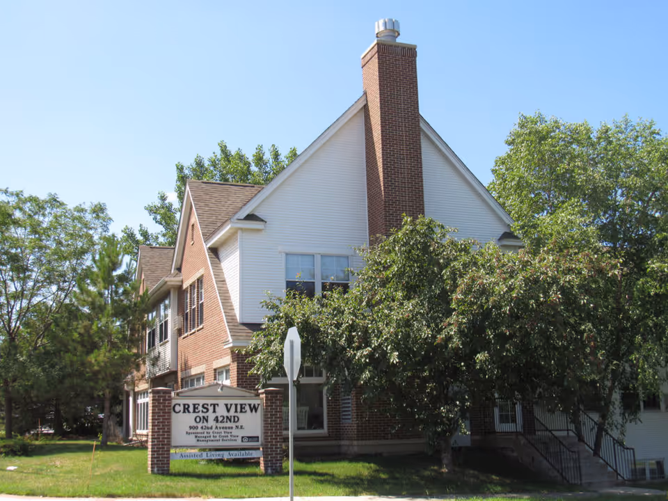 Exterior view of a two-story senior living facility named Crest View On 42nd, featuring a brick and white siding building with a prominent chimney, surrounded by trees and greenery under a clear blue sky. A sign in front displays the facility name and address.
