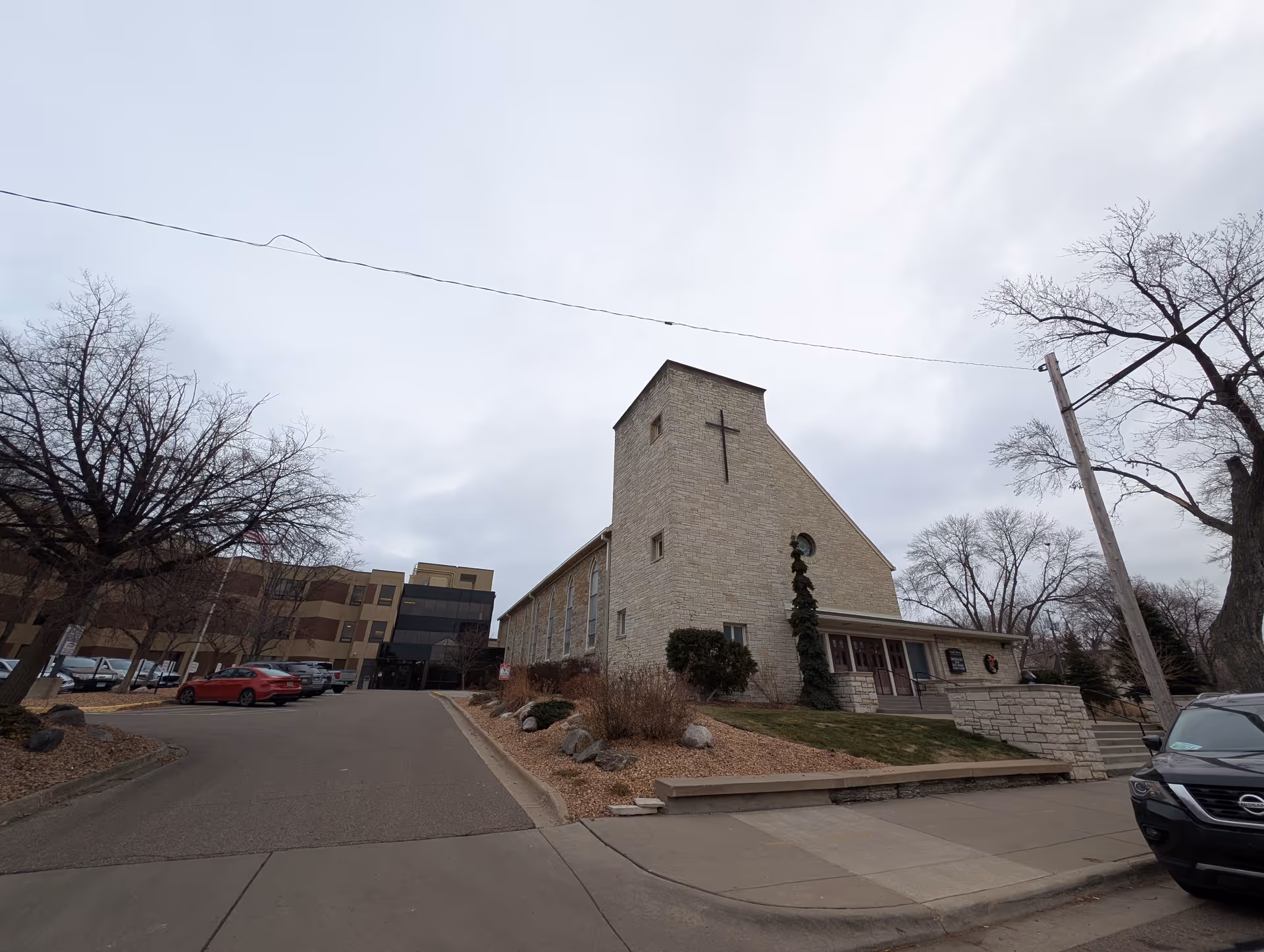 Exterior view of a senior living facility with a stone building featuring a large cross on the front, adjacent to a multi-story building with glass and beige panels. There are parked cars along the driveway and leafless trees around the property under a cloudy sky.