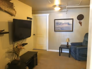 Small living room with a wall-mounted TV, blue recliner, side table with a lamp, framed artwork, and a closed door.
