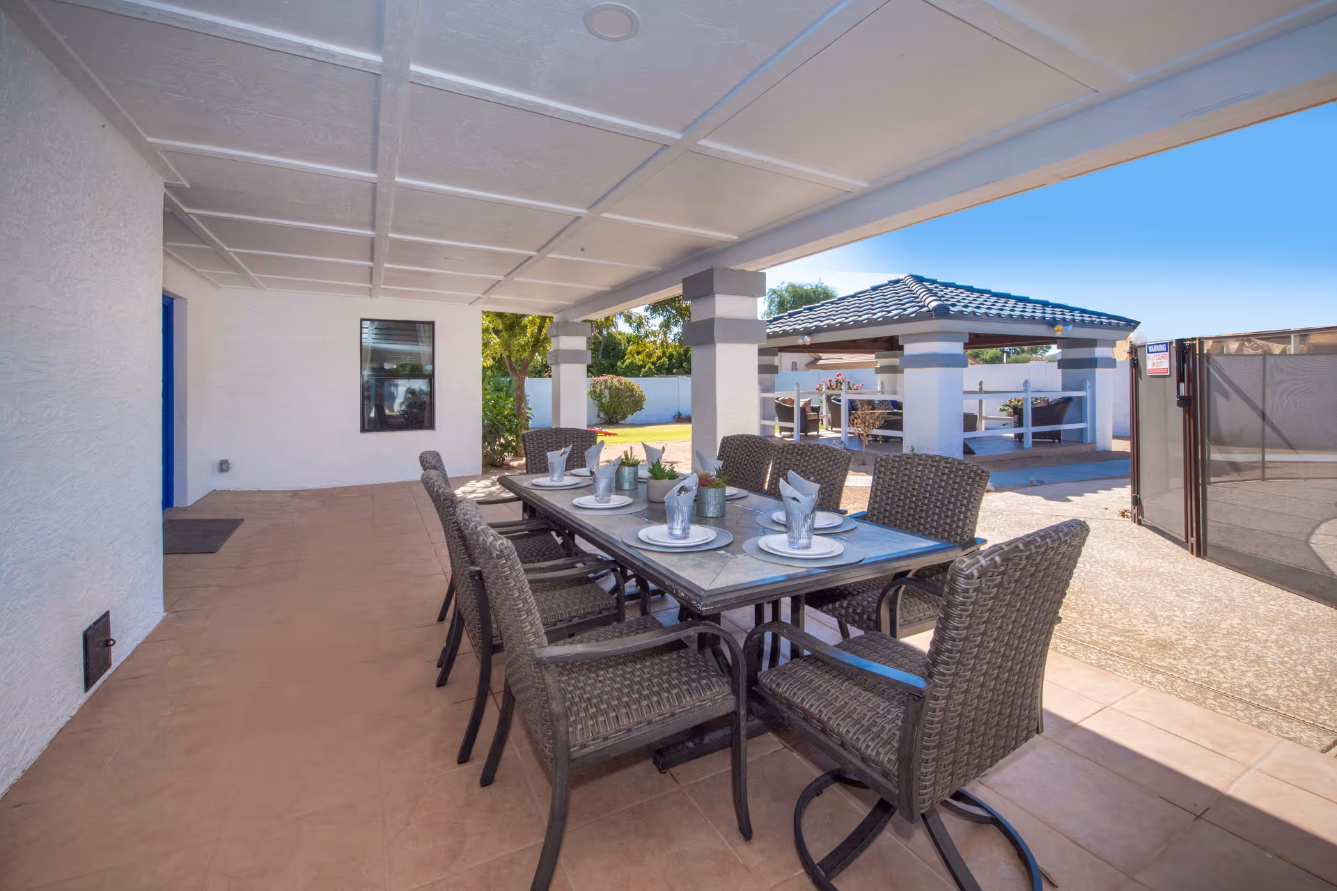 Covered outdoor patio with a long dining table set with plates and wicker chairs, facing a gazebo and courtyard.