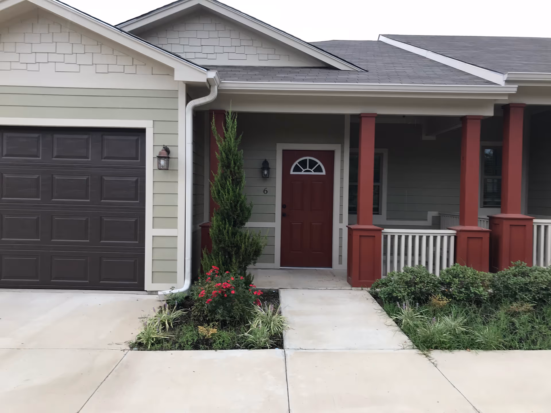 Front entrance of a single-story unit with a red door, attached garage, porch with red columns, and small landscaped beds.