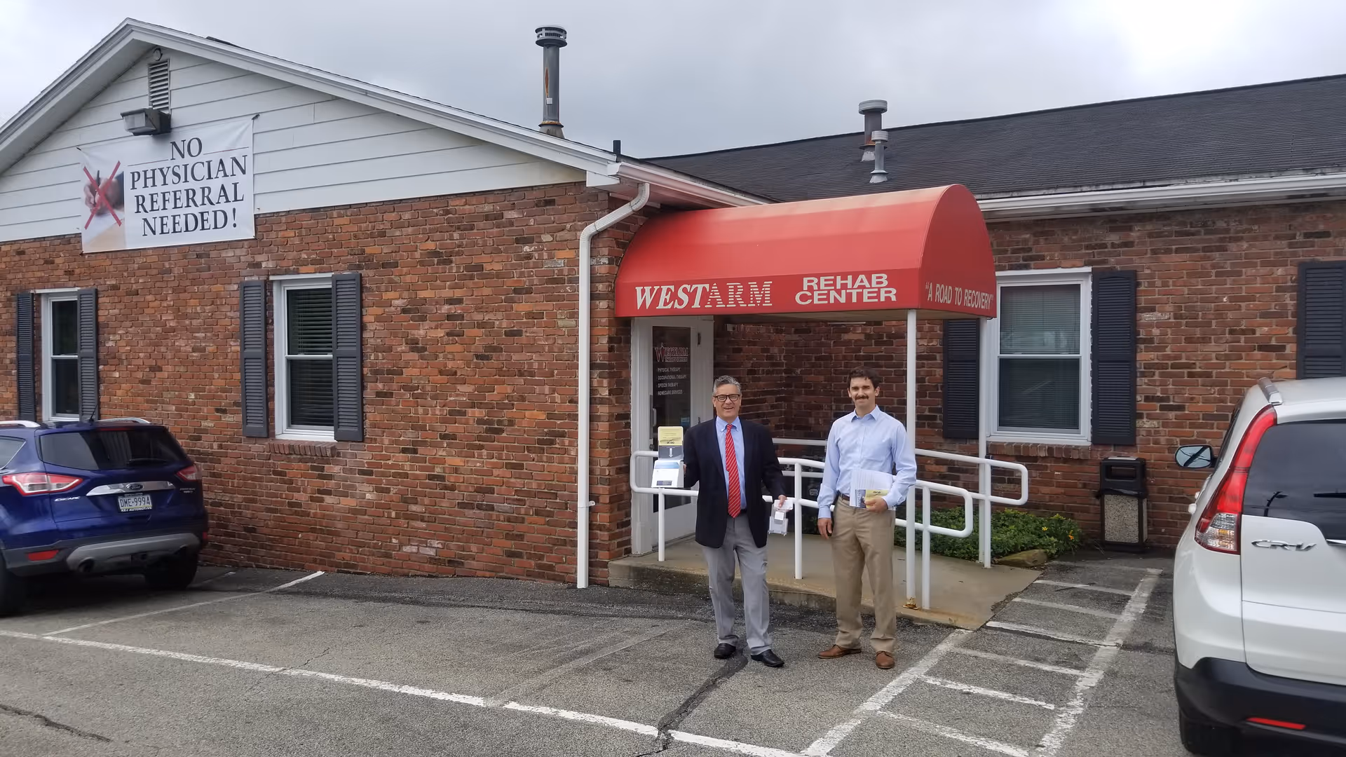 Brick rehab center exterior with red canopy, sign, and two men near the entrance.
