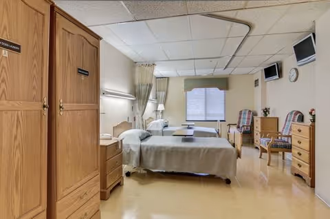 Shared patient bedroom with two beds, wooden dressers, curtains, and a window.