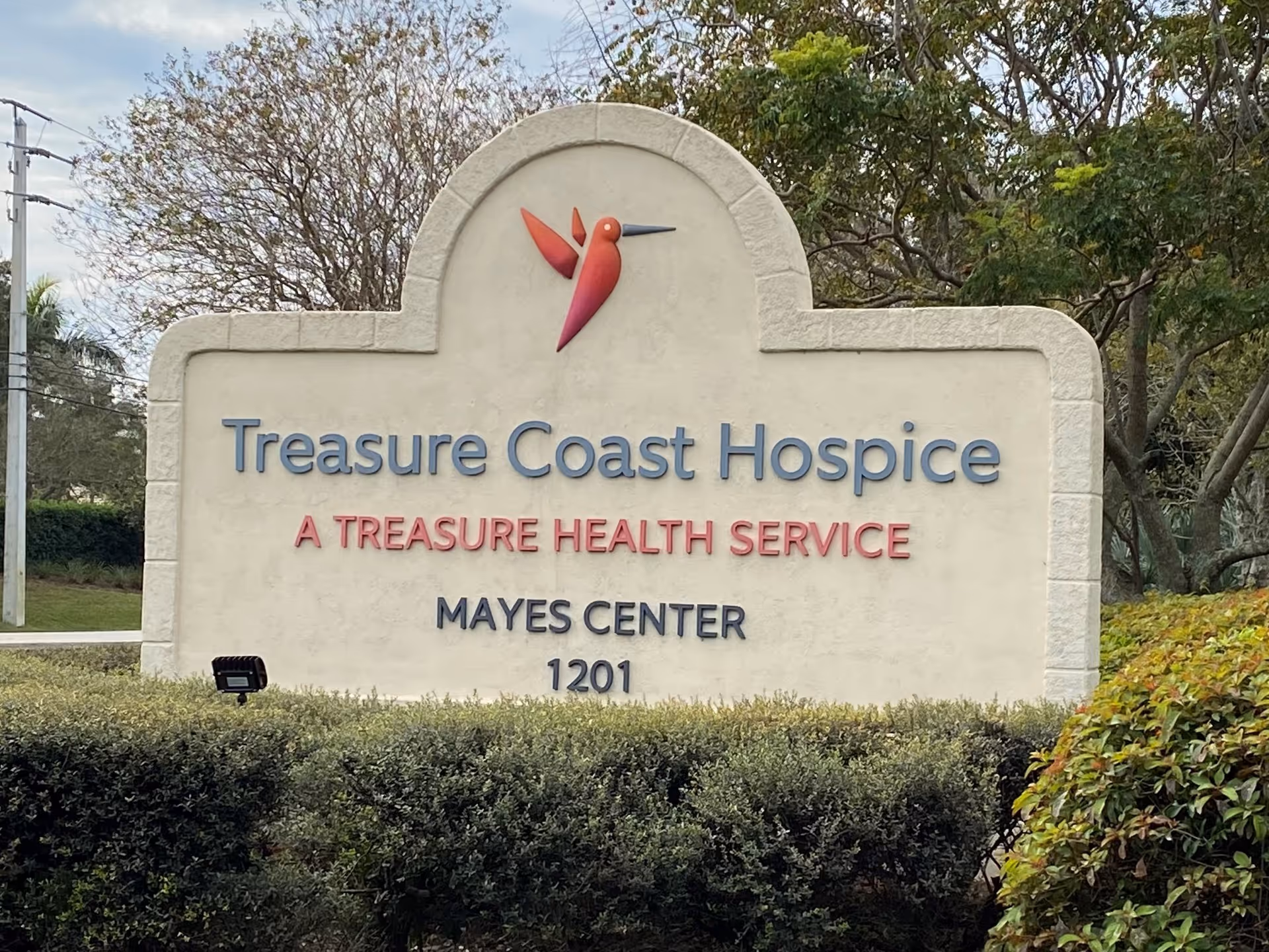 Large stone sign reading Treasure Coast Hospice with a red hummingbird logo, set in hedged landscape.