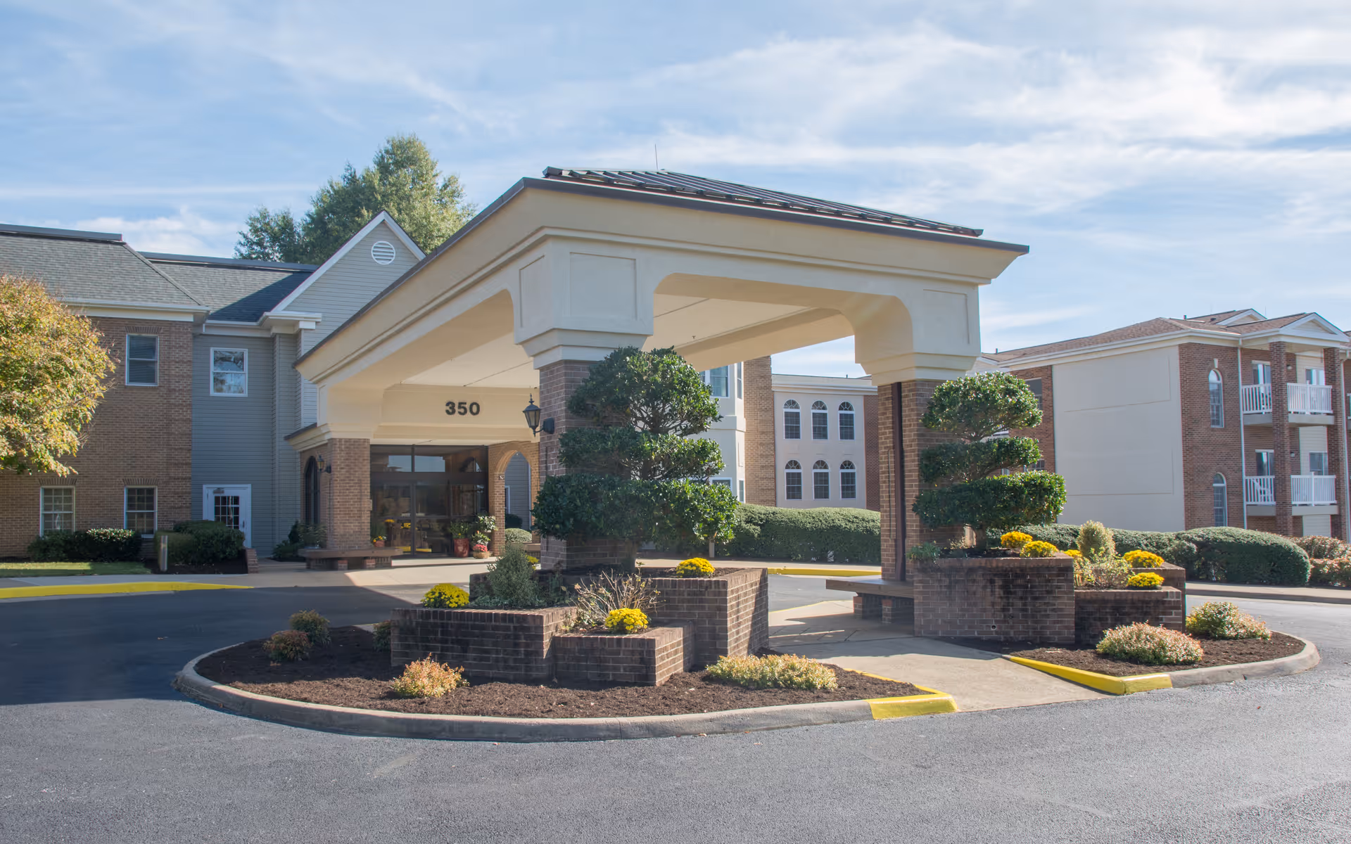 Entrance of King's Grant Retirement Community featuring a covered drop-off area with brick pillars and neatly trimmed bushes and flowers in landscaped beds under a partly cloudy sky.