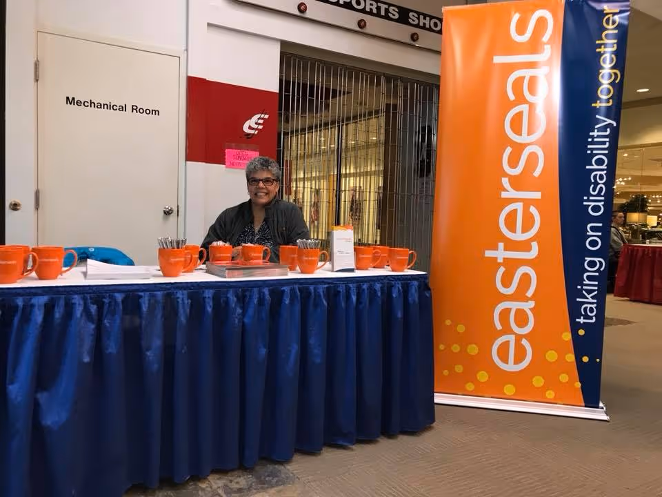 Smiling woman stands behind a blue booth table with orange mugs at Easterseals Alaska.