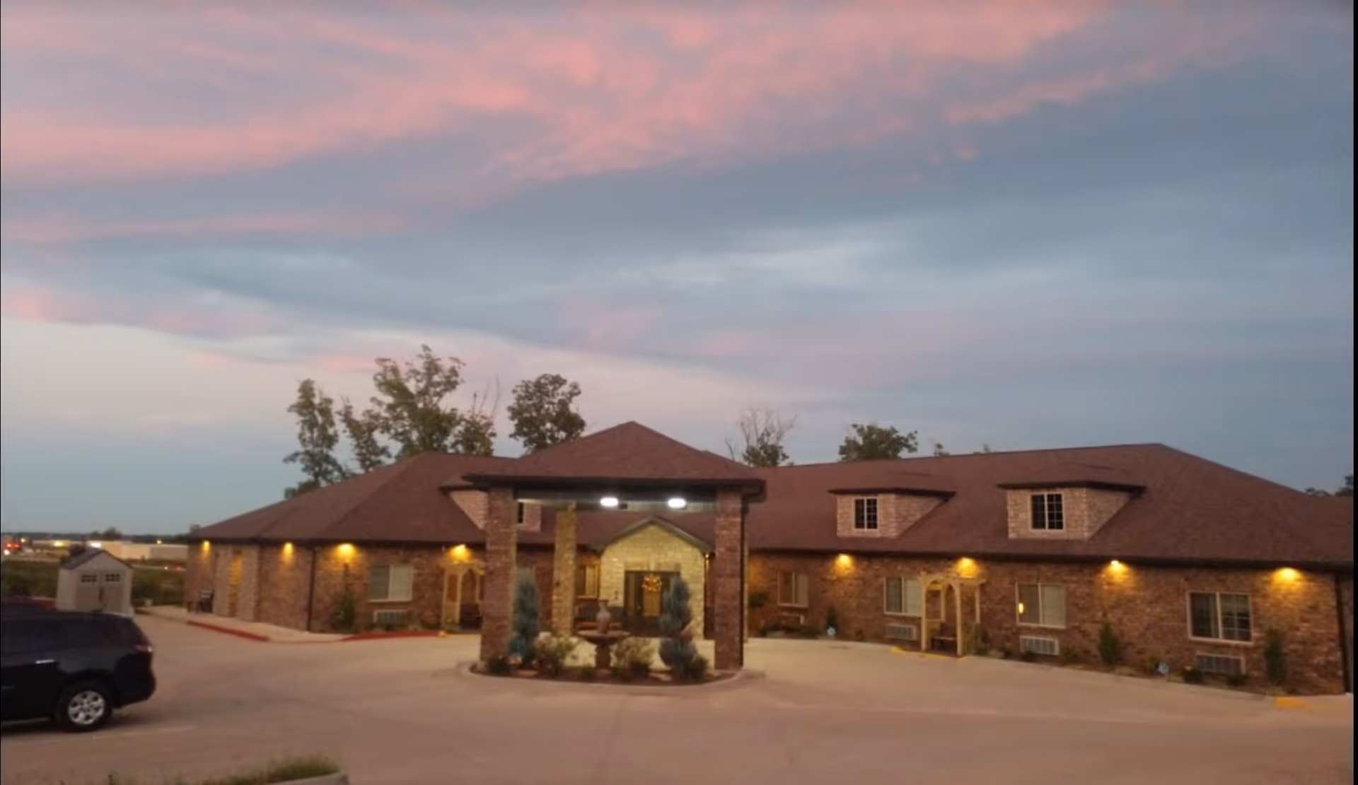 Exterior front view of a single-story brick senior living building at dusk with a lit entrance and driveway.