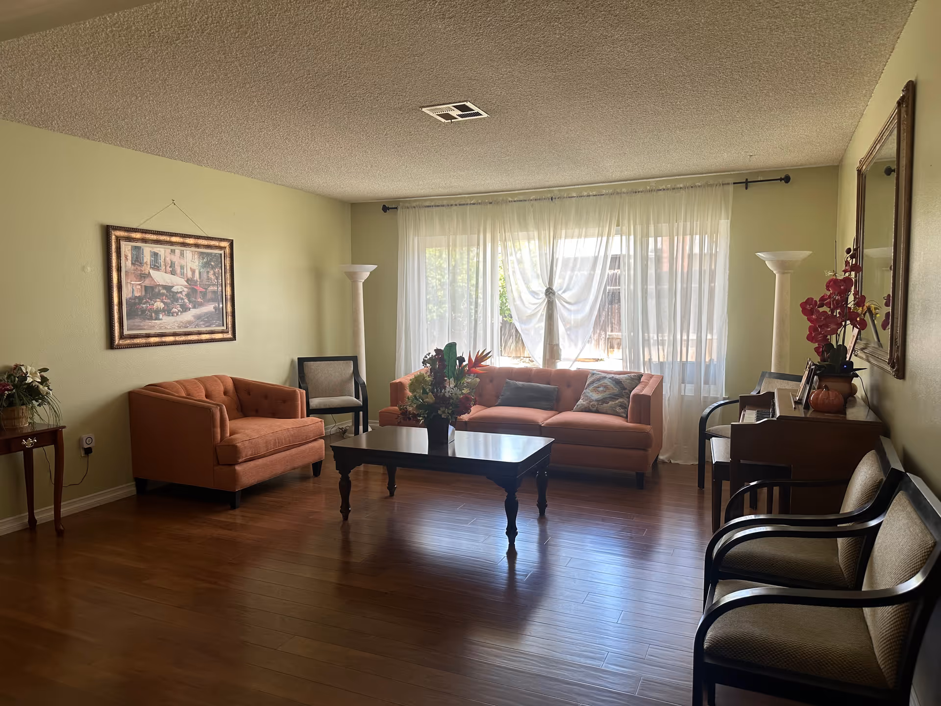 A cozy living room with wooden flooring, featuring two orange sofas, a dark wooden coffee table with a flower arrangement, two floor lamps, several chairs, a side table with a plant, and a large window with sheer white curtains allowing natural light to fill the room. There is a framed painting on one wall and a large mirror on the opposite wall.