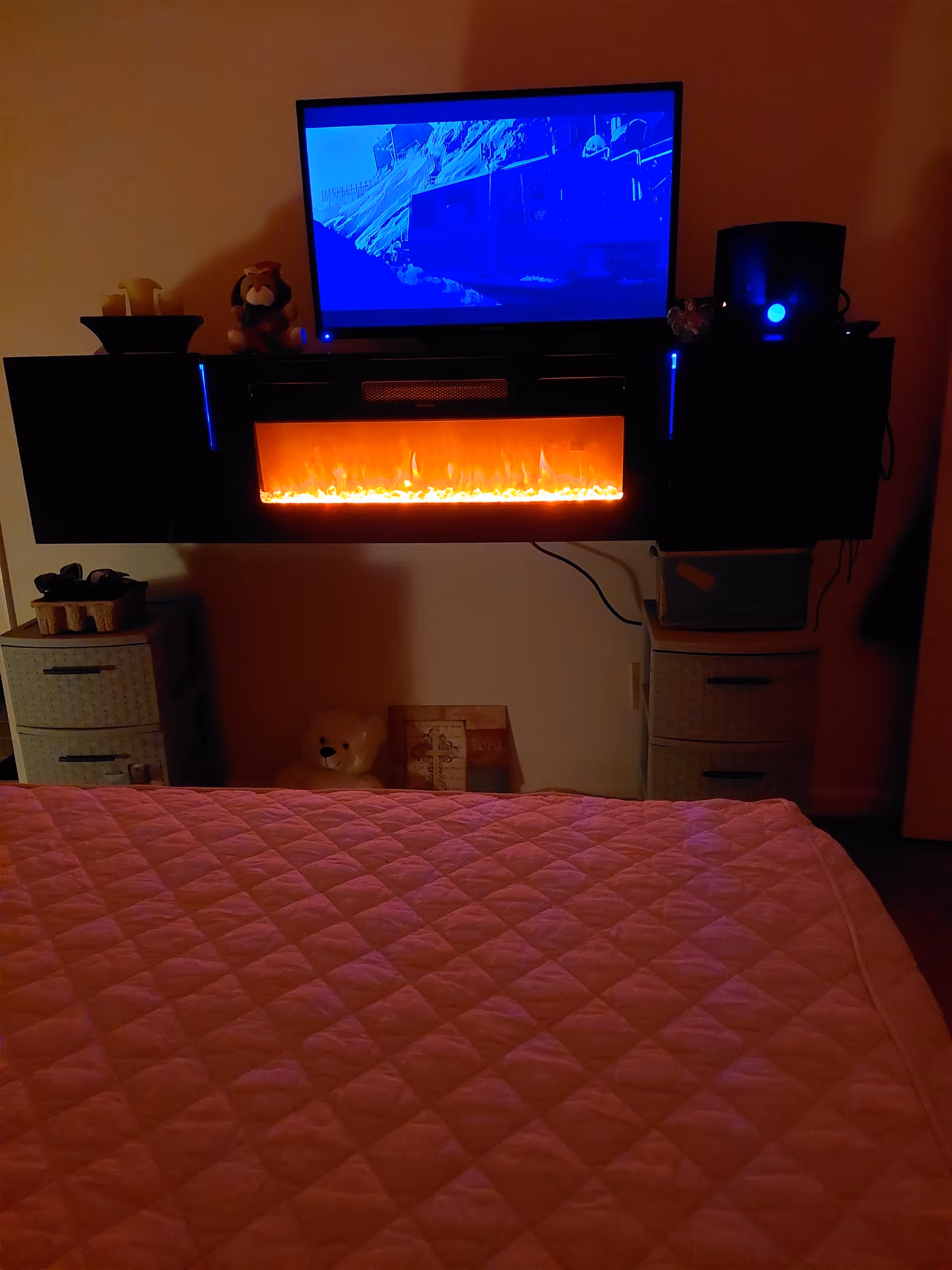 Bedroom scene with a pink quilted bedspread, mounted TV, electric fireplace, and bedside drawers.