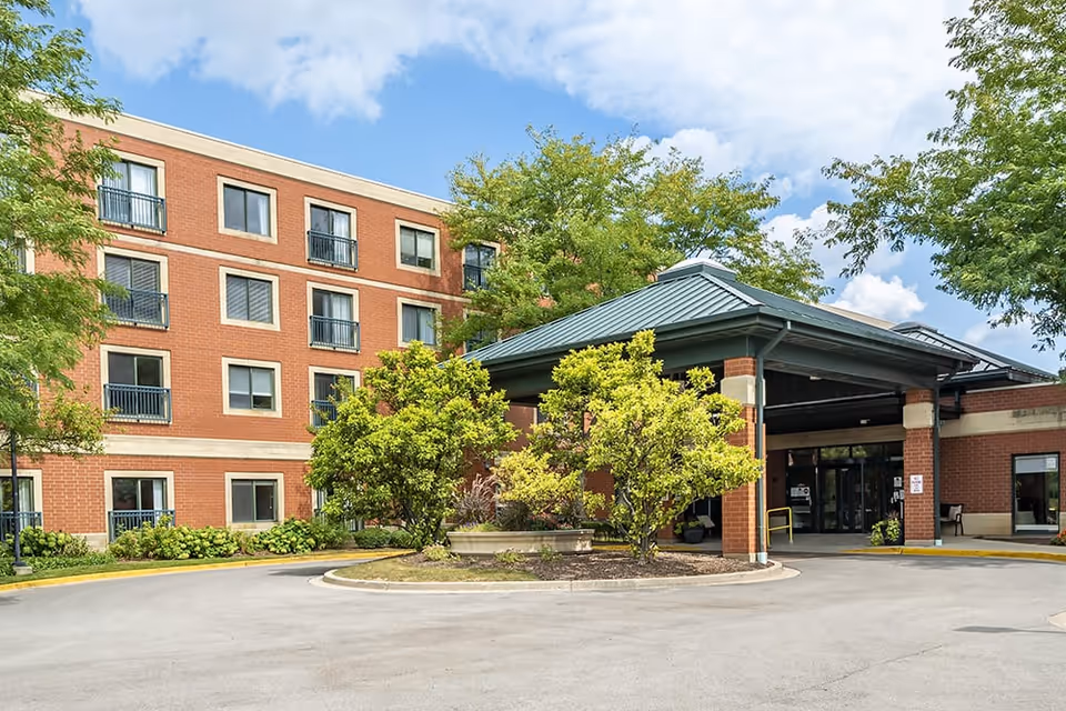Front entrance of a brick senior living building with a covered porte-cochère, trees, and landscaping.