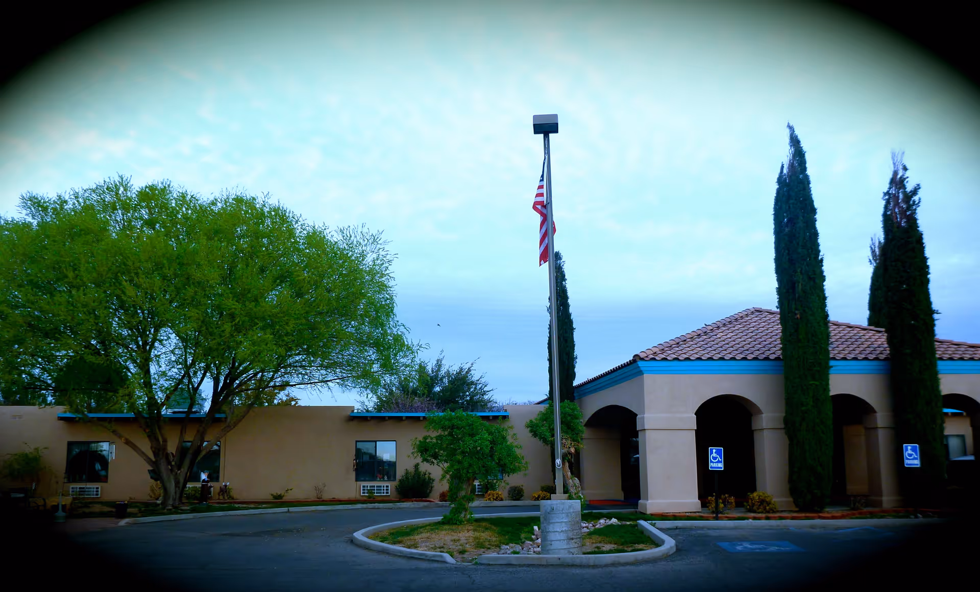 Front exterior of a single-story assisted living building with a flagpole, trees, tiled roof entryway, and handicap parking signs.