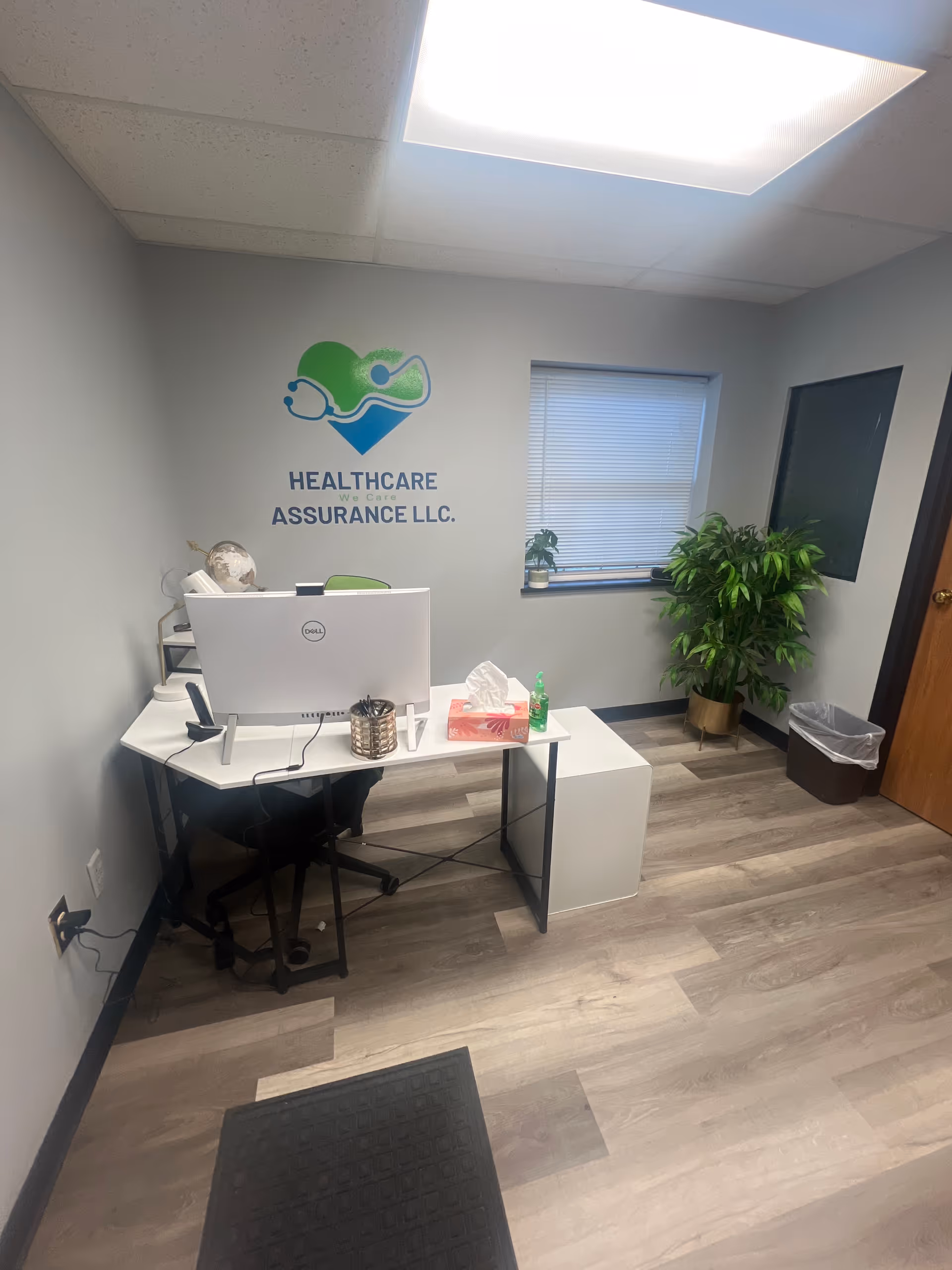 Office workspace with a computer desk, plants, and HealthCare Assurance LLC logo on the wall.
