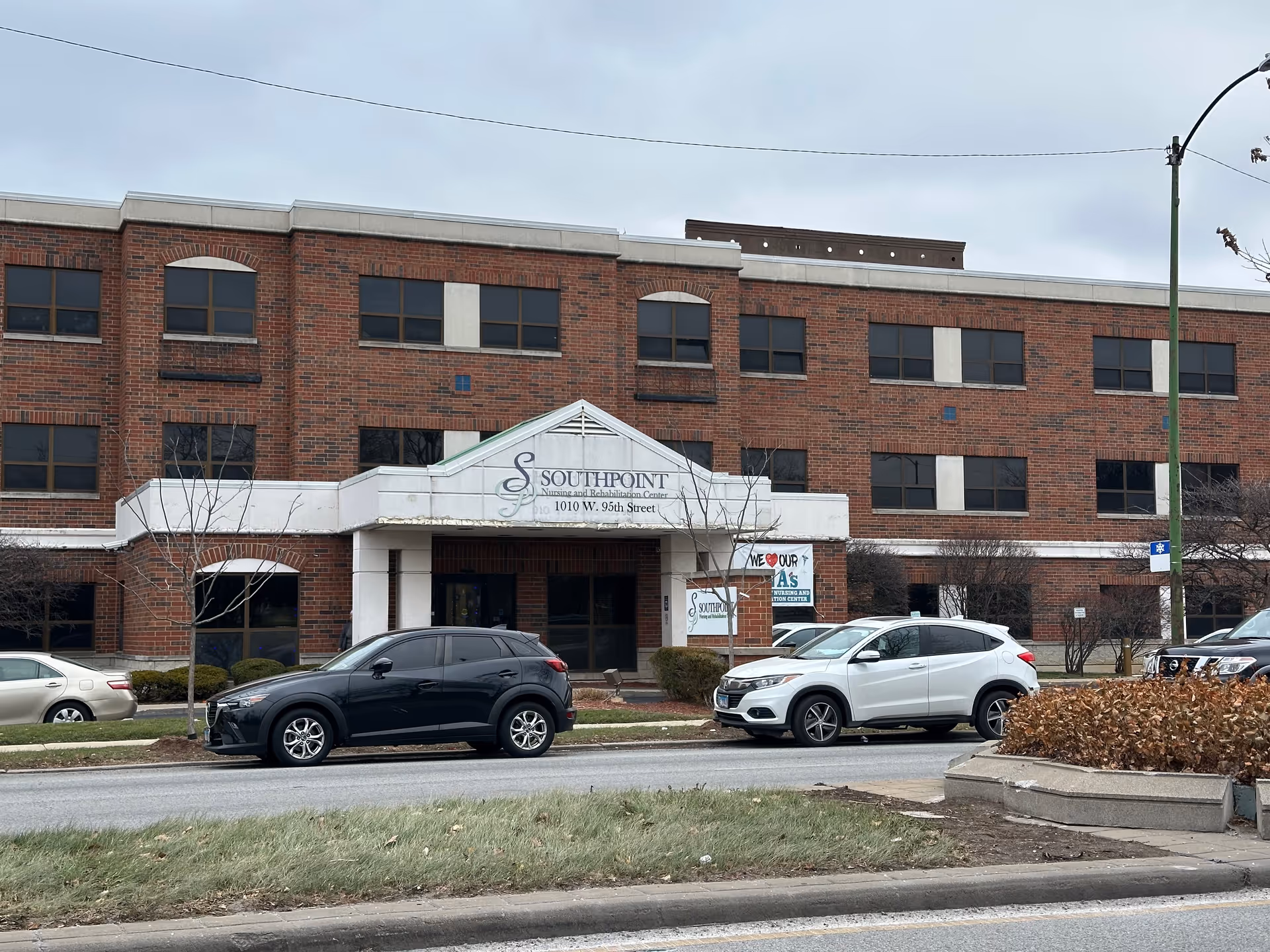 Front exterior of the Southpoint Nursing and Rehabilitation Center, a three-story brick building with a covered entrance and cars parked in front.