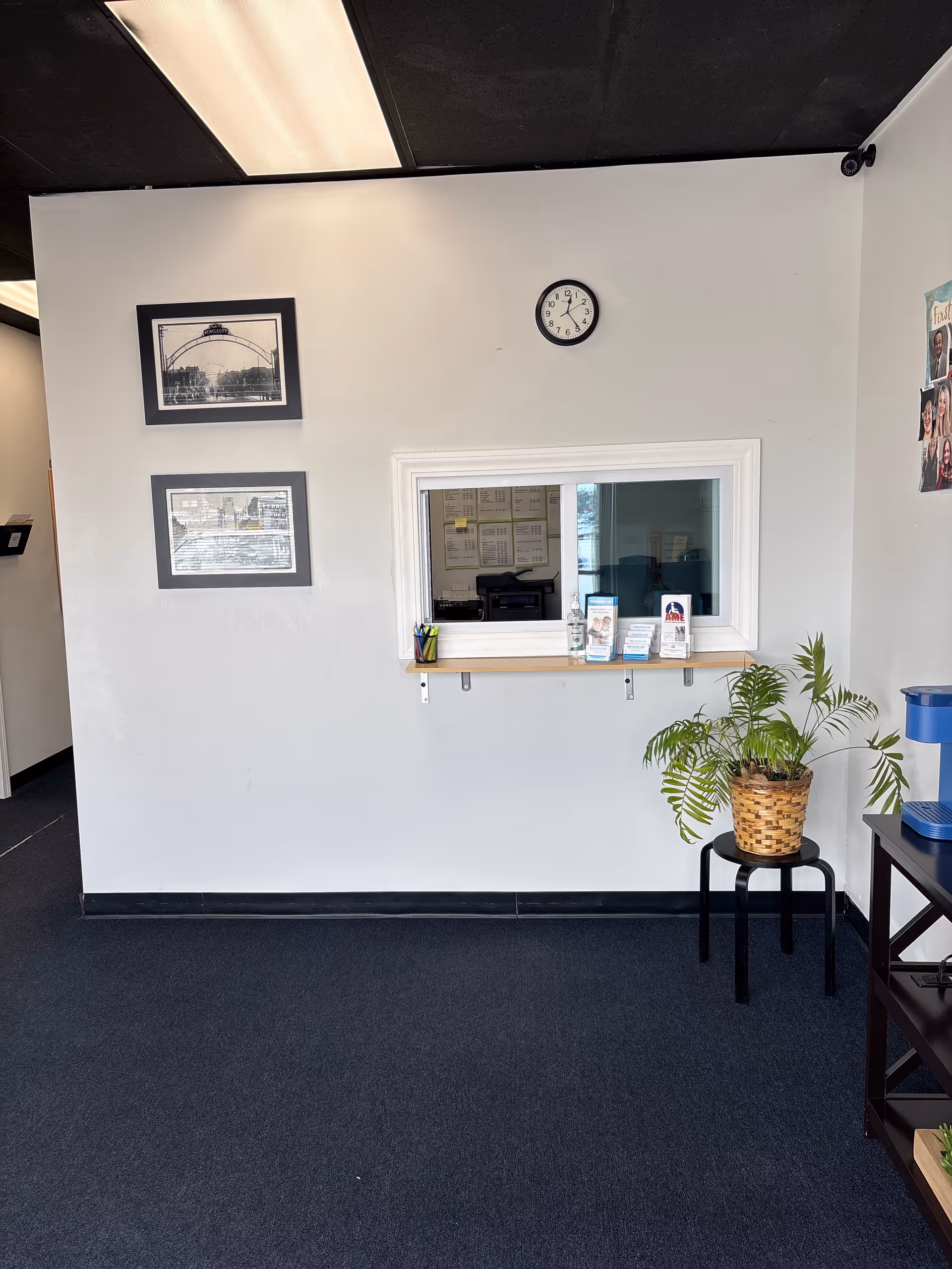 Reception window in a white wall with a clock, plant, and framed photos.