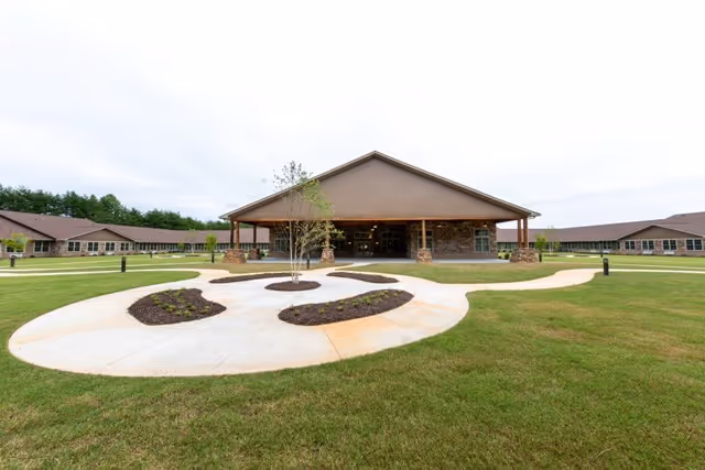 Wide exterior view of a single-story assisted living facility with a large covered entrance, surrounded by well-maintained green lawns and a curved concrete walkway with small landscaped planting beds.