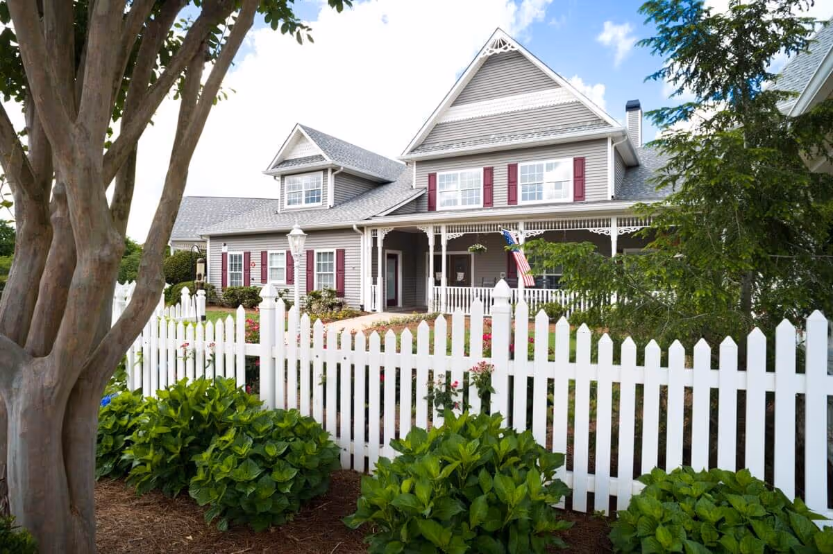 A large two-story house with gray siding and red shutters behind a white picket fence. The house has a covered porch with decorative trim and an American flag hanging near the entrance. There are green bushes and trees in the garden surrounding the fence, and a clear blue sky with some clouds in the background.