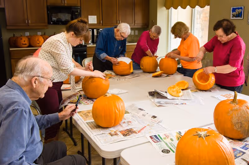 A group of elderly people and a younger woman gathered around a large table indoors, carving and decorating pumpkins. The table is covered with newspapers and several pumpkins, some carved and some whole, are visible. The room has wooden cabinets and a window with a yellow valance.