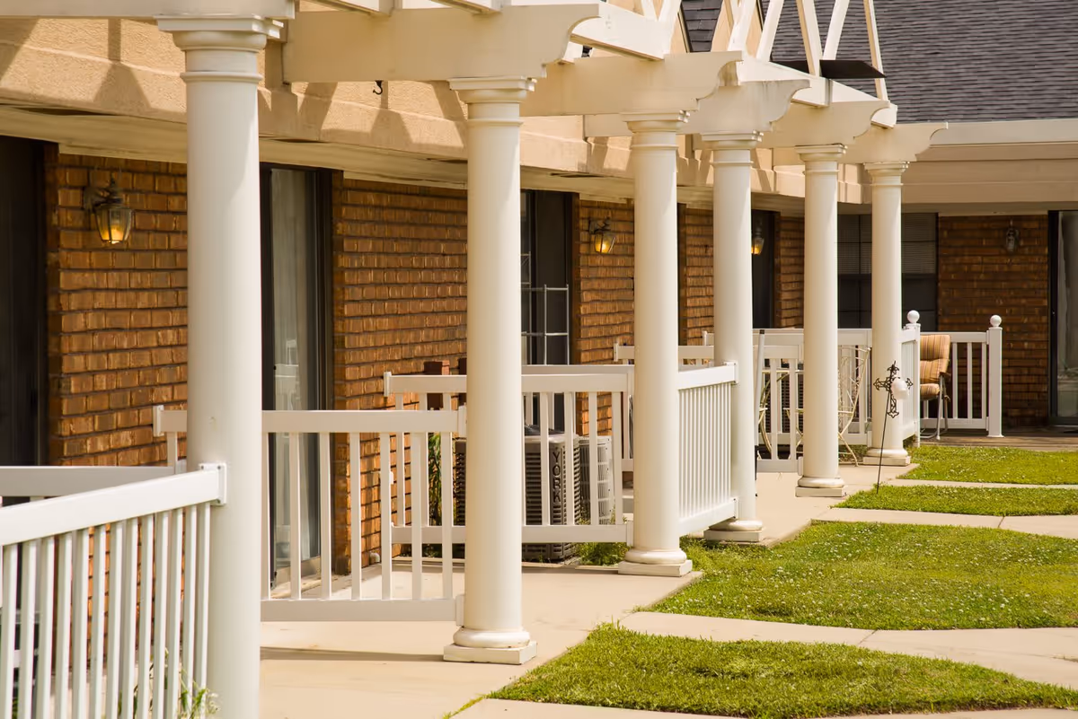 A row of white columns and porches along the brick exterior of an assisted living building with grass and paved walkways.