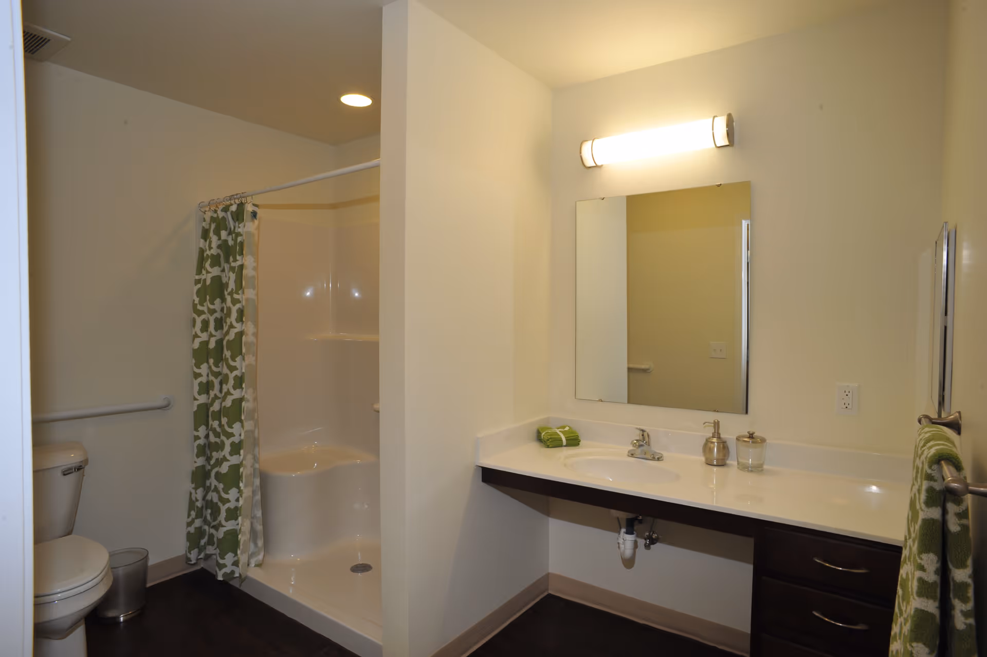 A clean and modern bathroom featuring a white toilet, a walk-in shower with a green and white patterned curtain, a large mirror above a white countertop with a sink, and a wall-mounted light fixture. There are green towels on the countertop and hanging on a towel rack.