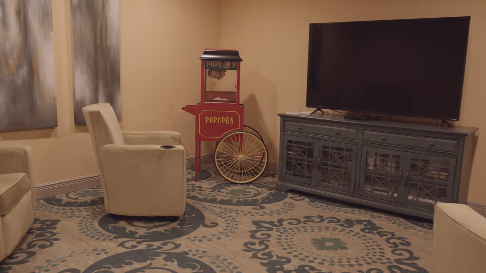 A cozy lounge area featuring two beige armchairs, a vintage popcorn machine in red, and a large flat-screen TV mounted on the wall. The floor is decorated with an intricate patterned rug.