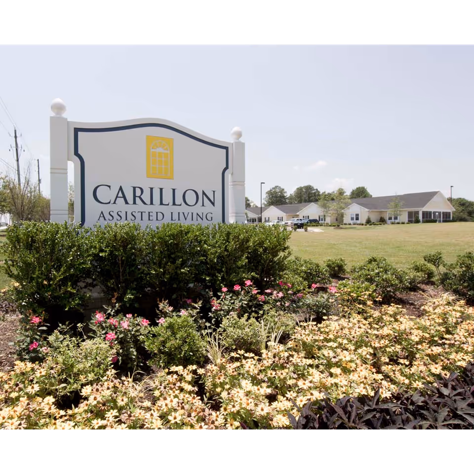 A large Carillon Assisted Living sign sits amid flowering landscaping with the facility buildings and lawn visible behind it.