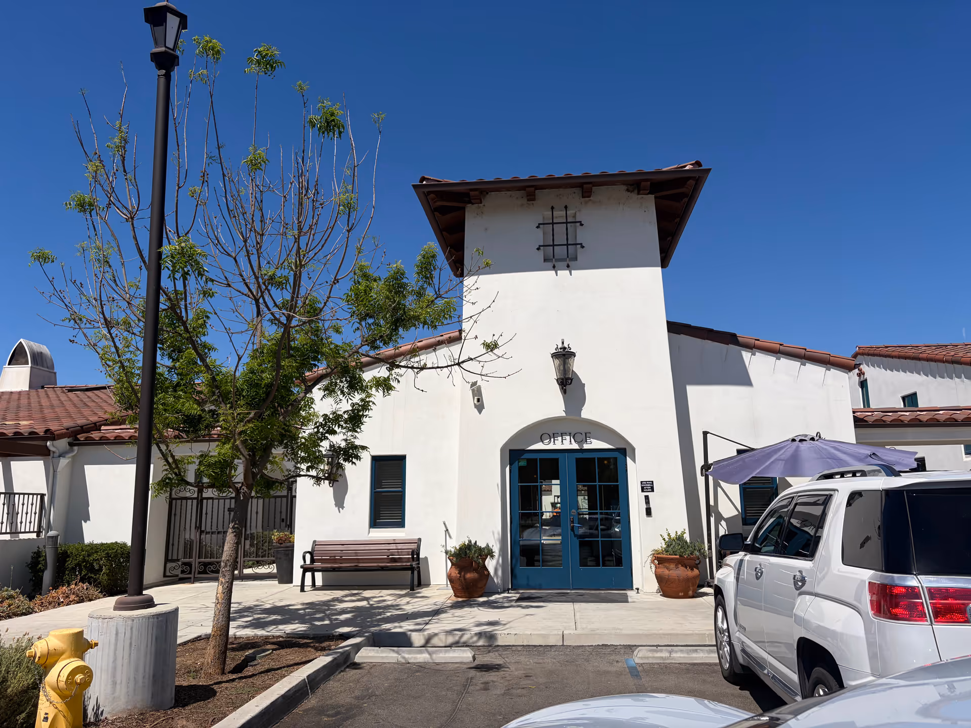 Office entrance of The Artesian of Ojai with blue doors, bench, trees, and a parked SUV.