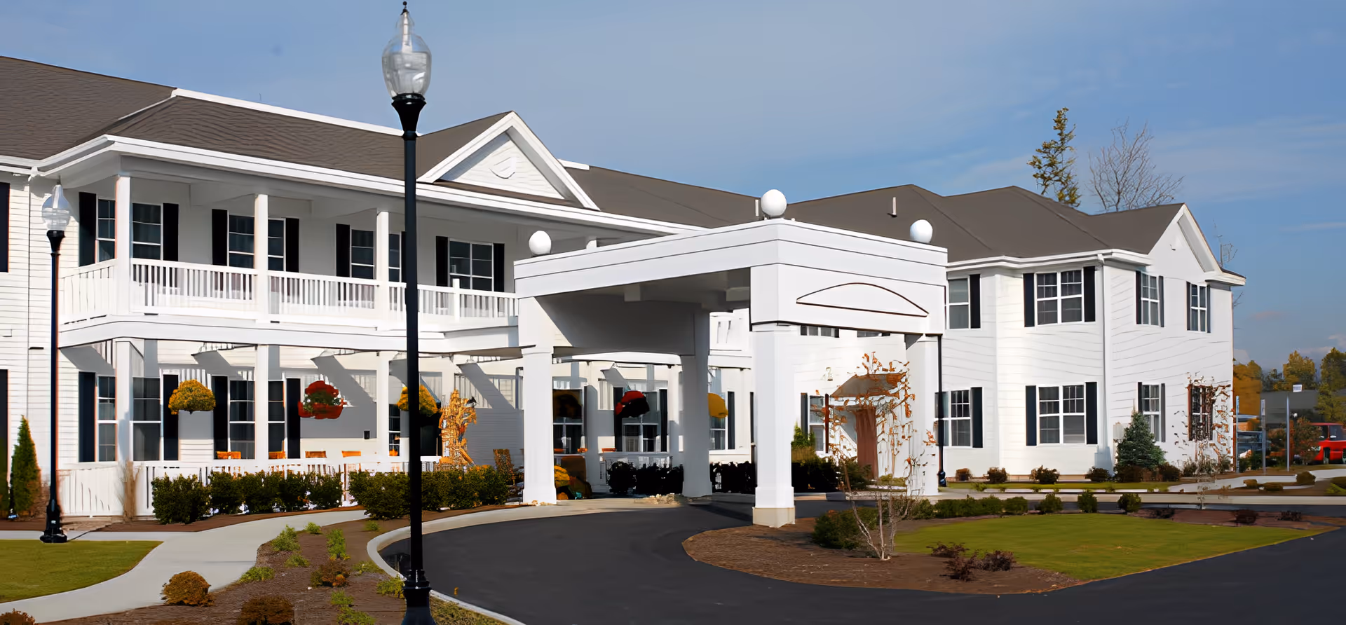 Exterior view of a large white senior living facility building with black window shutters, a covered entrance with columns, landscaped grounds, and a clear blue sky.