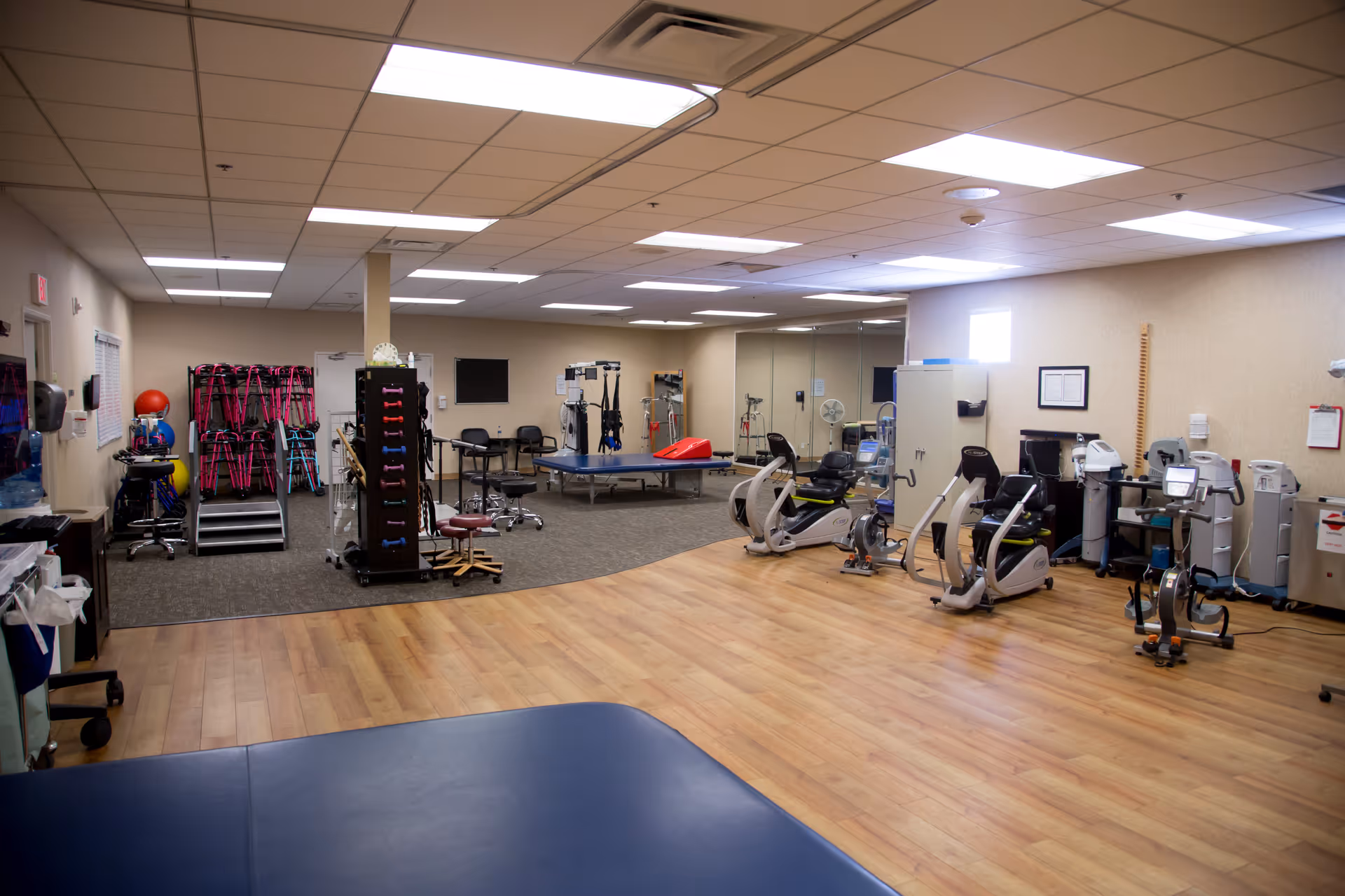 Bright therapy and exercise room with recumbent bikes, treatment tables, and various rehabilitation equipment and storage racks.