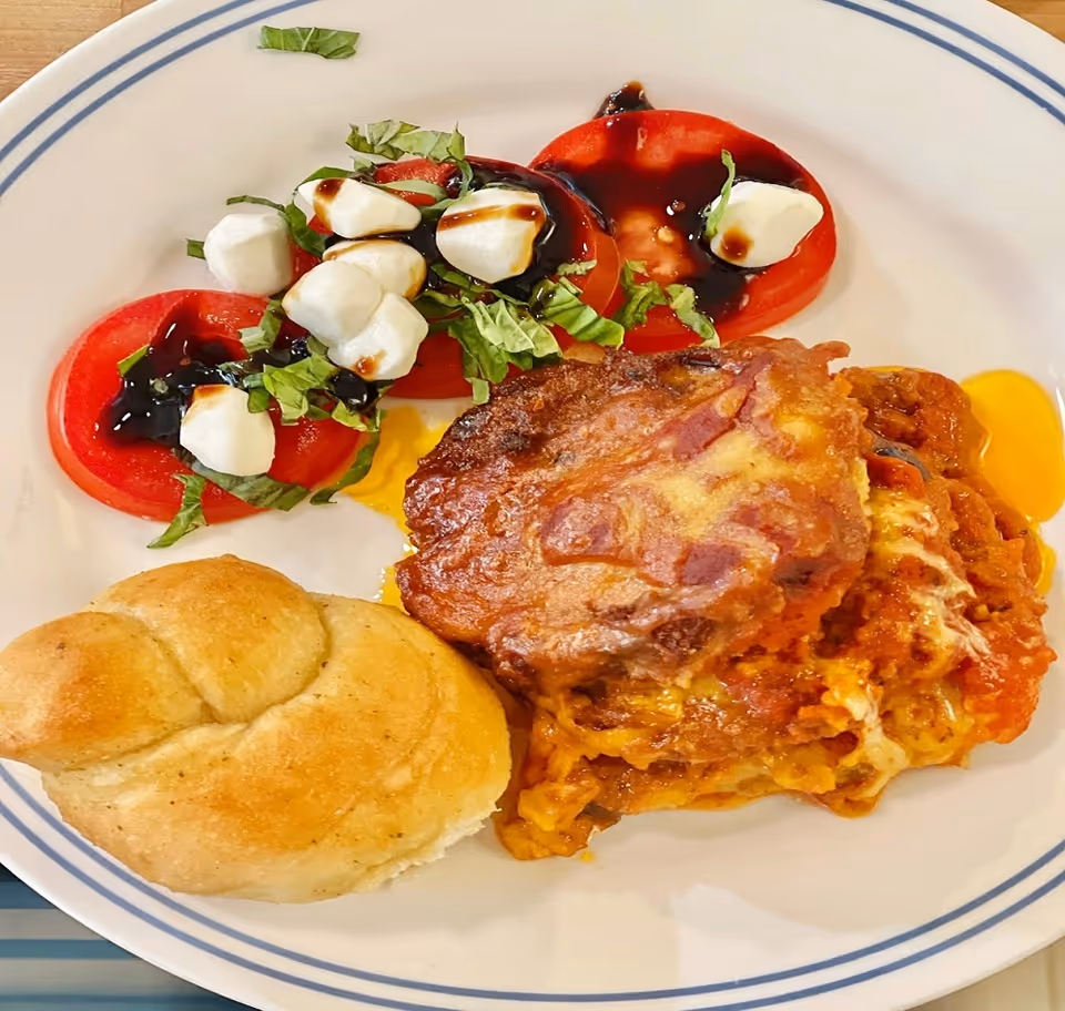 Plate features tomato slices with mozzarella, greens, balsamic, plus a cheesy meat casserole and roll.