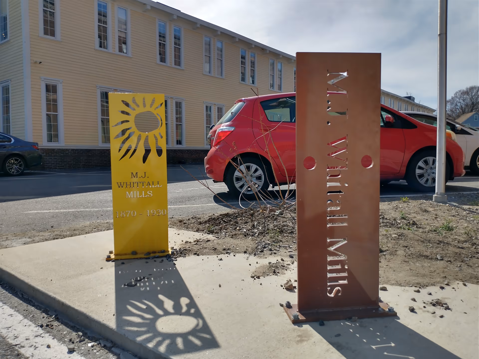 Exterior view of Embrace Midwifery facility with yellow sun sign, rusted vertical panel, and parked cars.