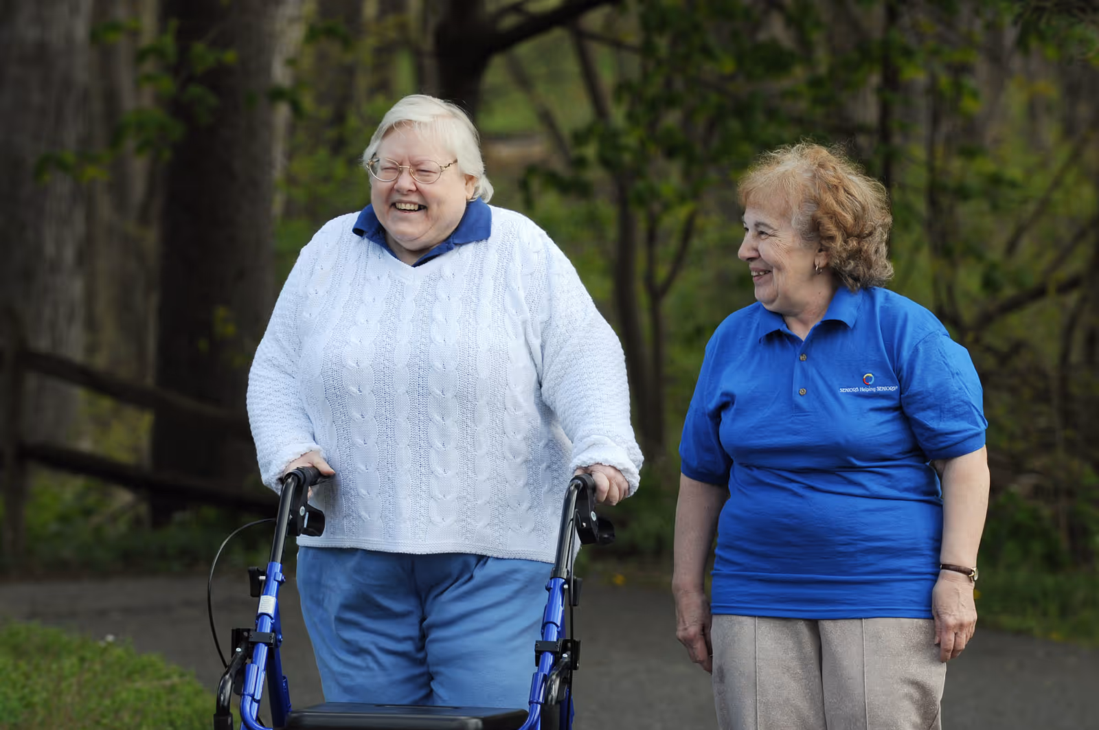 Two smiling seniors walk outdoors on a park path, one using a walker.