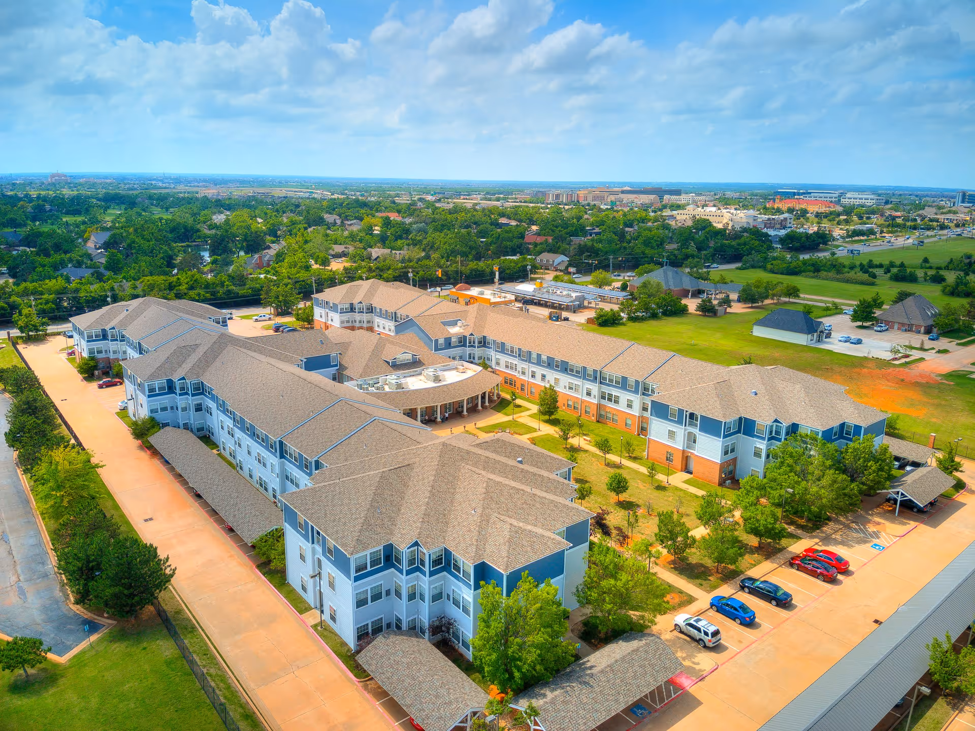 Aerial view of Town Village senior living facility showing multiple connected buildings with beige roofs and blue and white exterior walls, surrounded by green lawns, trees, and parking areas with several cars. The background includes a suburban landscape with more buildings and greenery under a partly cloudy sky.