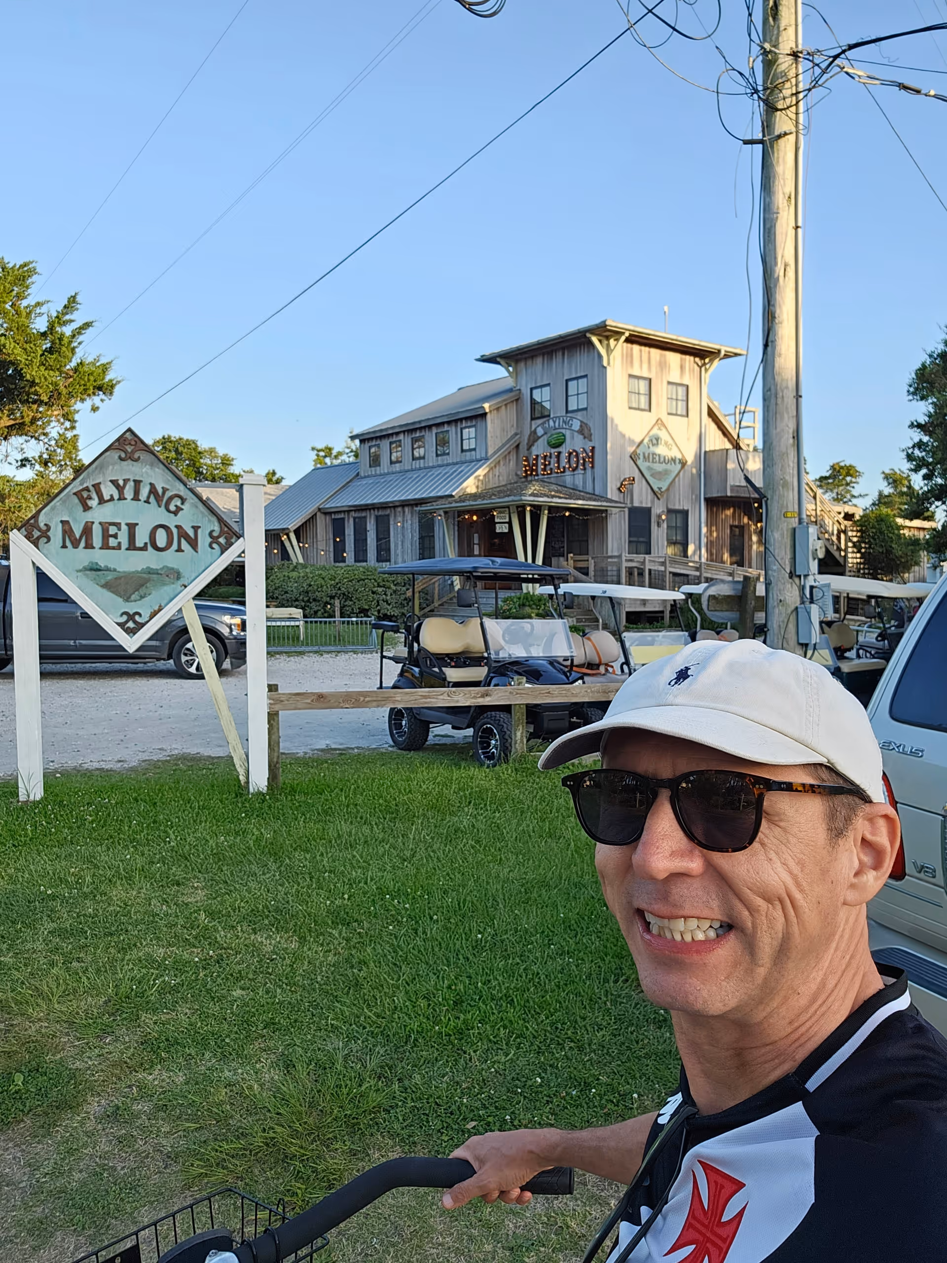Man wearing sunglasses smiles near rustic Flying Melon building and golf carts outside.