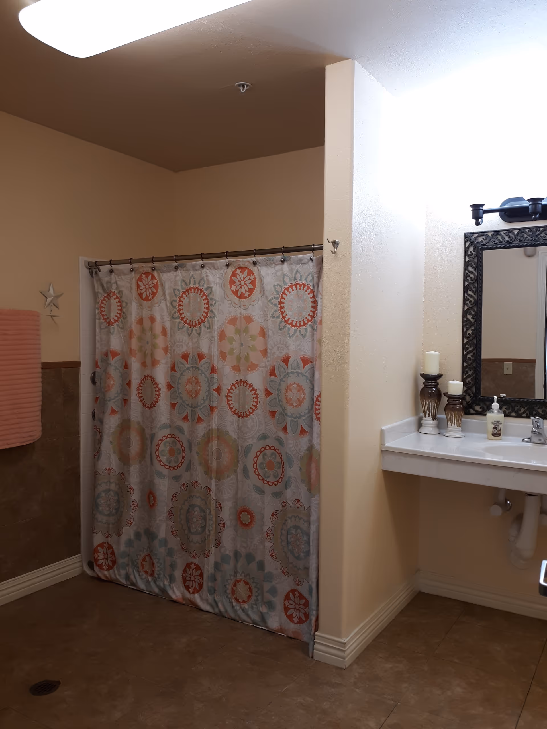 Narrow bathroom with patterned shower curtain, sink, mirror, candles, and beige walls.