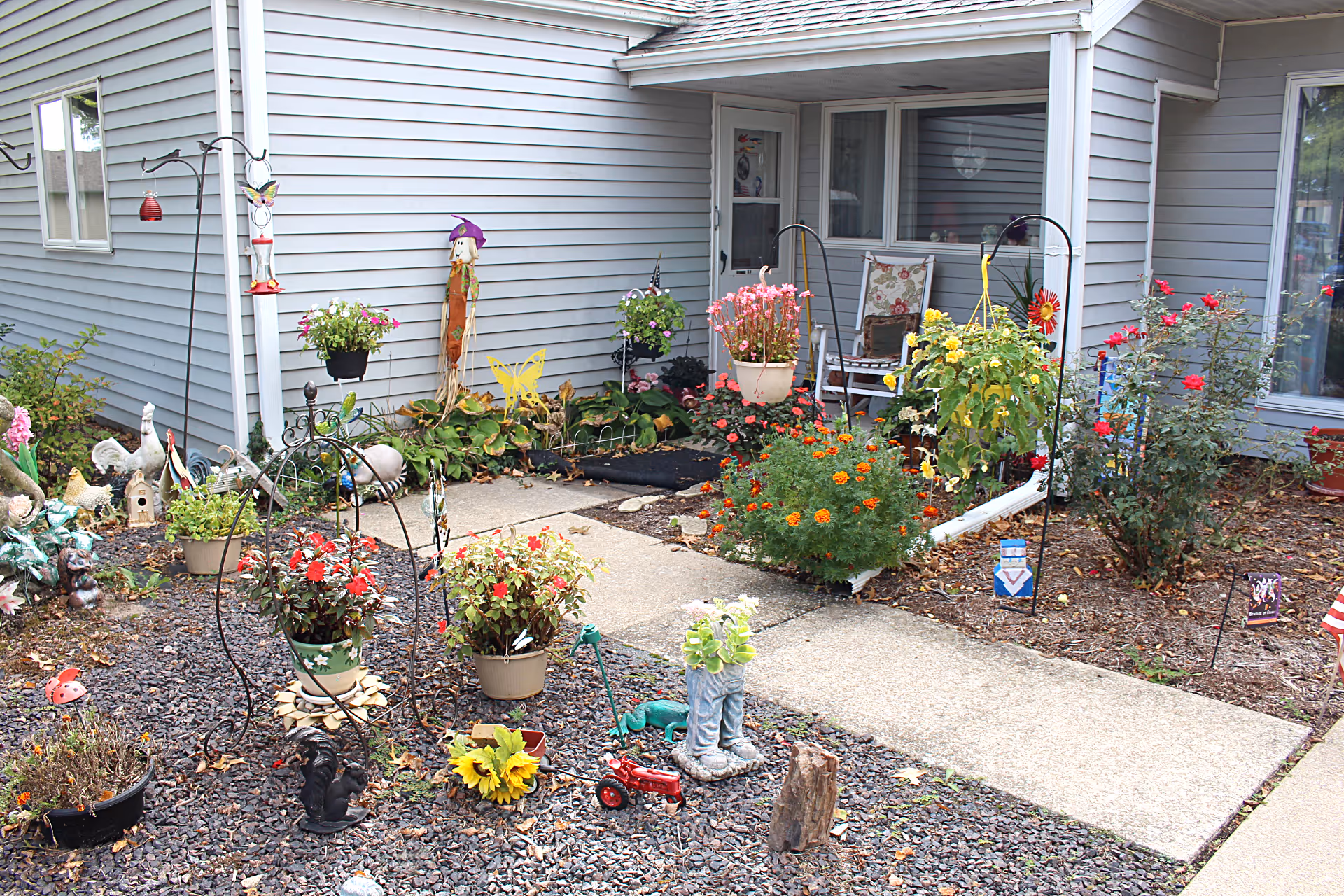 Entrance to a gray-sided building with a small front garden filled with potted flowers, decorative ornaments, and a concrete walkway.